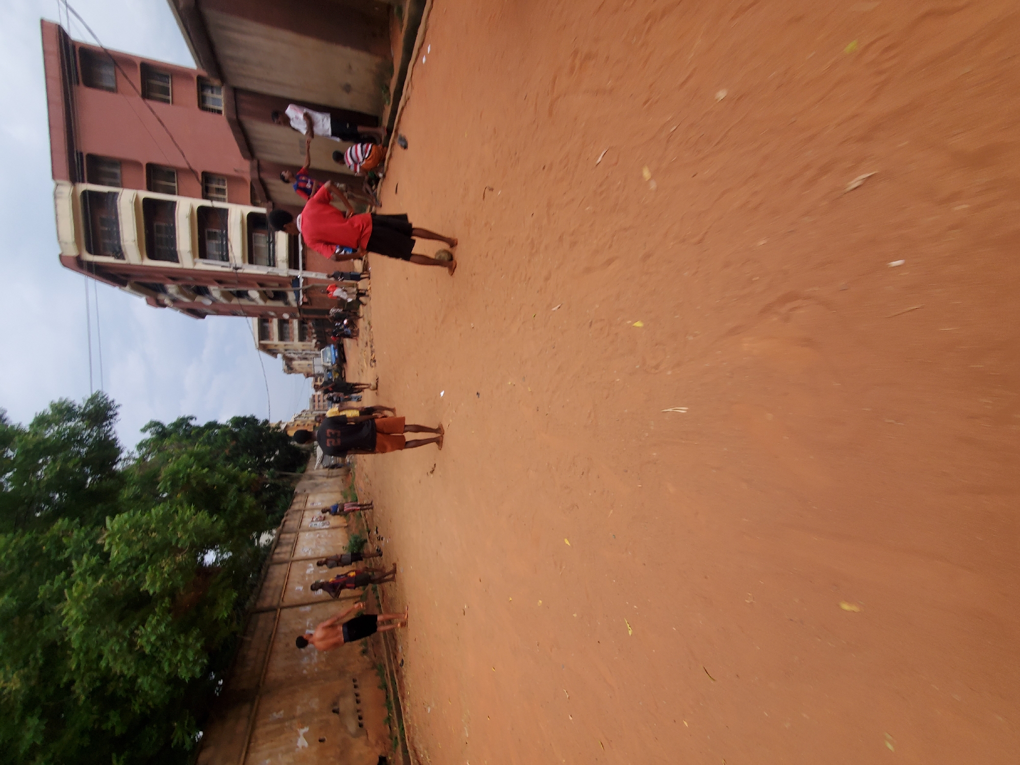 The image depicts a street scene in Onitsha, Nigeria. The ground is red earth. A group of people are walking along the street, with some performing exercises near a wall. Buildings are visible on one side of the street, and trees on the other. There are power lines overhead.