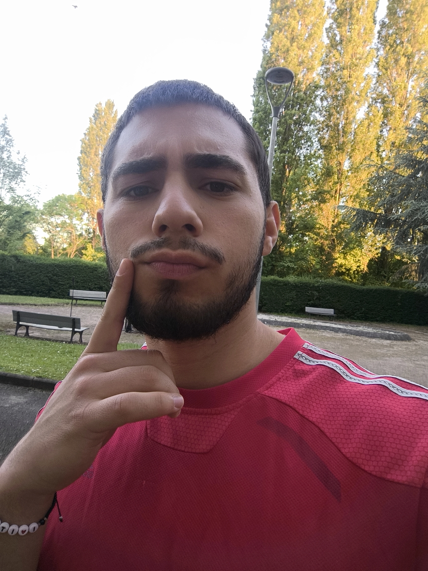 A medium shot depicts a man with short dark hair, a beard, and wearing a red athletic shirt with white stripes on the shoulders. He is holding his finger to his chin as if deep in thought. The background reveals a park setting with benches, a lamp post, manicured hedges, and tall trees, indicating an outdoor location. Based on the known associations, the park is in Schiltigheim, France.