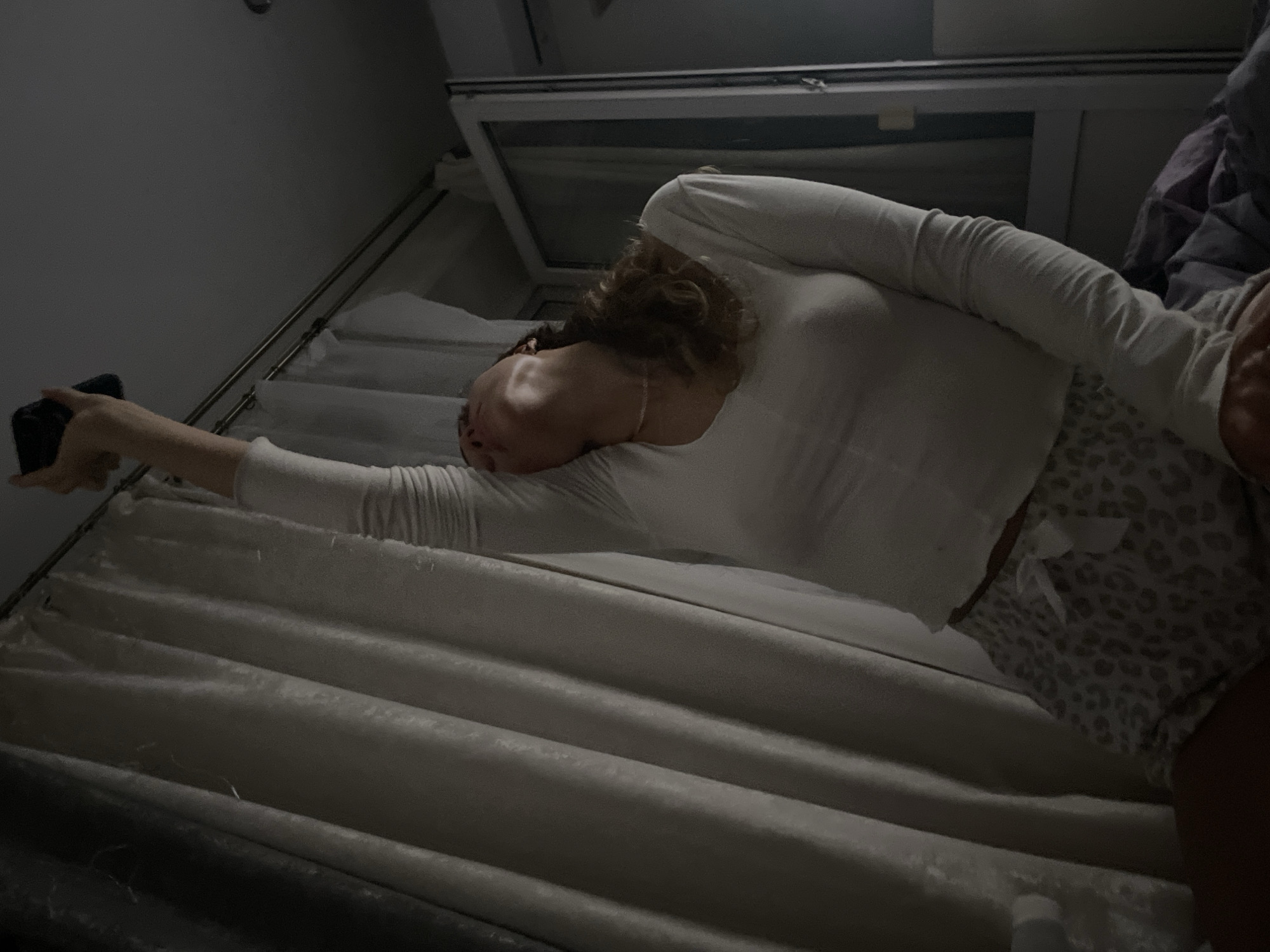 An upward-angled, dimly lit indoor photograph captures a young individual, possibly female, reclining on a bed in a room in Unknown, Unknown. The individual has light skin, curly light brown hair, and wears a long-sleeved white top and patterned light-colored shorts with a white bow. A thin, light-colored necklace is visible around her neck. Her head is tilted backward, exposing her neck, as her right arm is extended vertically upward, grasping a dark-colored smartphone or similar small electronic device.
To the left of the individual, cream or light beige heavy curtains are visible, hanging from a metallic rod with rings, covering a significant portion of a window or wall. The curtains exhibit folds and texture. Above the curtains and the individual, a dark, featureless ceiling is visible. To the right of the individual, a white-framed window or balcony door is partially open, revealing a dark exterior or another room beyond. A rumpled purple and gray blanket or duvet covers part of the bed beneath the individual. The overall lighting suggests a nighttime or early morning setting, with ambient light illuminating the scene from an unseen source.