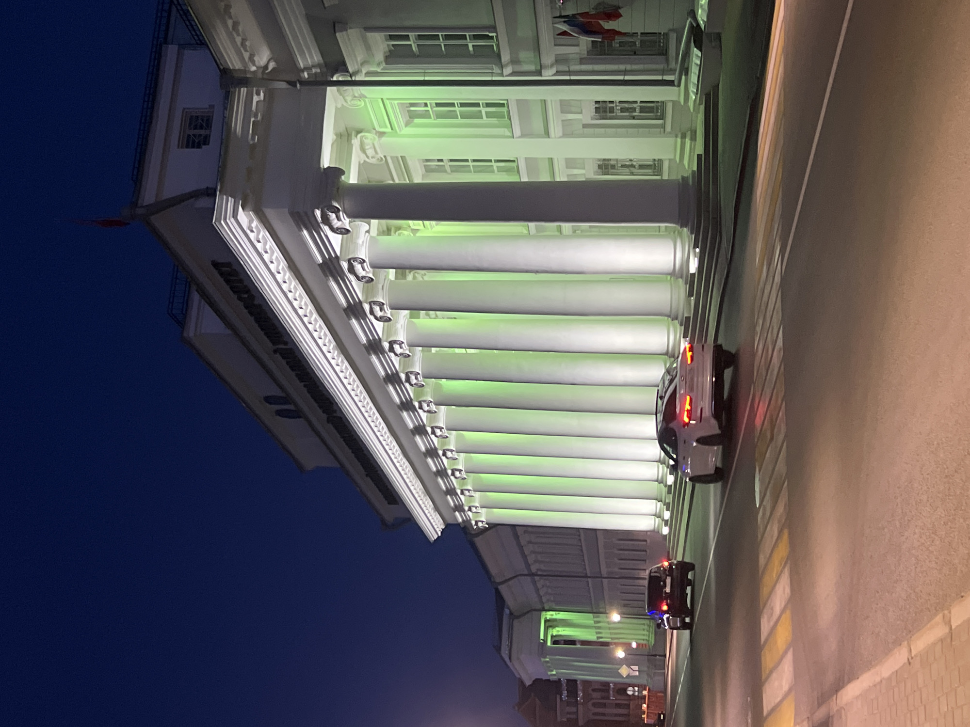 The image depicts a building with a long row of white columns lit with green light, located in Kazan, Russia. A white car and a black car drive on the road next to the building. The sky is dark blue, suggesting it is nighttime. The road has a marked pedestrian crossing. A flag is visible on the building's roof. There are lights on the street near the end of the building.