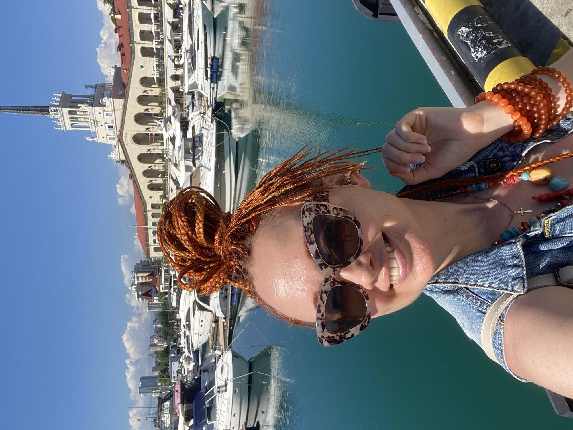 A woman with bright orange braided hair styled in a bun is taking a selfie in a marina in Sochi, Russia. She is wearing a denim jacket, sunglasses with a tortoise-shell pattern, and multiple necklaces and bracelets. In the background, numerous yachts are docked in the turquoise water, and a large, ornate building with a tall tower, identified as the Sochi Sea Port, is visible. The sky is clear blue with scattered white clouds. The woman is smiling and looking at the camera, holding one of her braids.