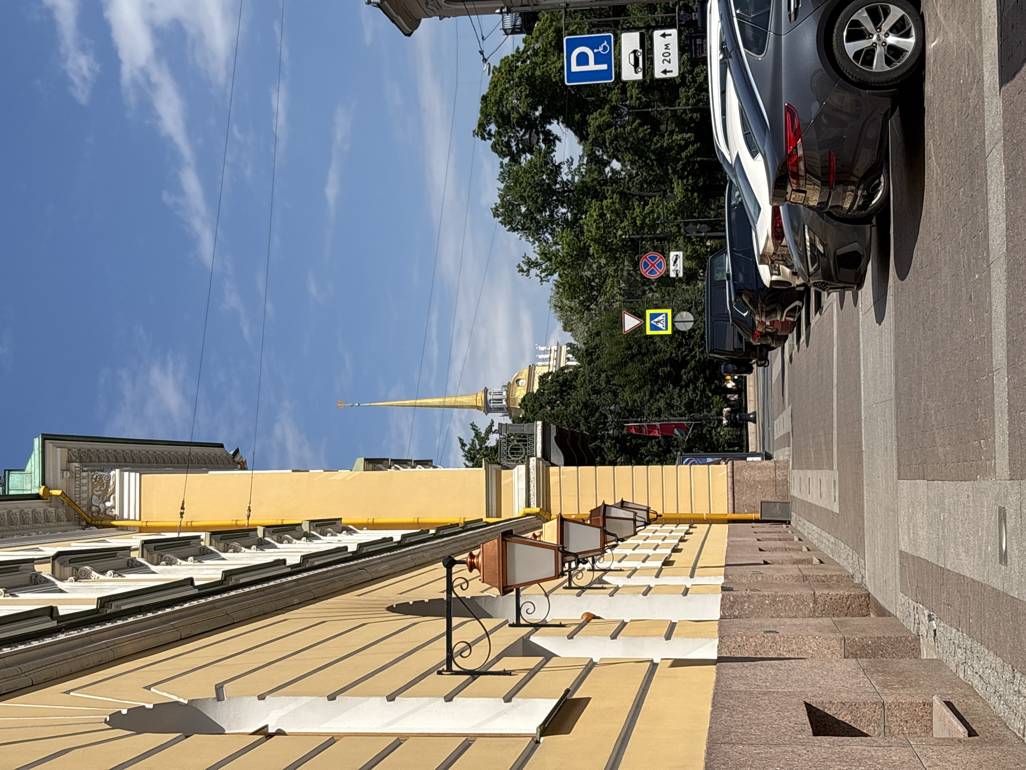 The image shows a street scene in Saint Petersburg, Russia, with a yellow building on the left and parked cars on the right. The building features elaborate architectural details, including decorative trim and lanterns mounted on the facade. In the background, a tall, golden spire, characteristic of Russian Orthodox churches, pierces the clear blue sky. Several traffic signs are visible, including a parking sign with a wheelchair symbol and a sign indicating "20m". The street is paved with cobblestones and lined with trees. The overall impression is of a well-maintained urban environment with historical architecture.