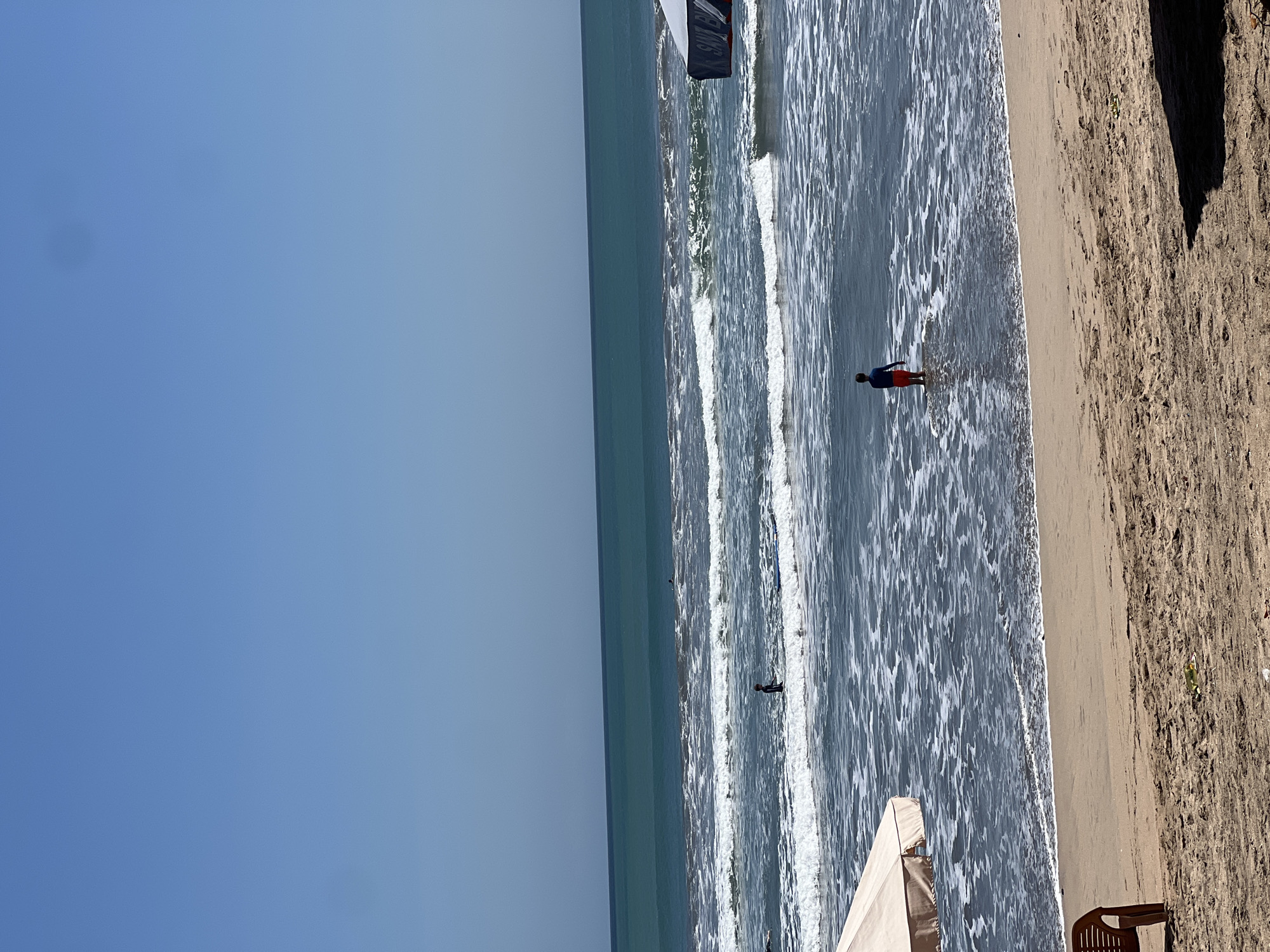 The image depicts a wide shot of Kuta Beach in Kuta, Indonesia, on a clear, sunny day with a bright blue sky. The ocean stretches out to the horizon, with turquoise water near the shore transitioning to a deeper blue further out. Gentle waves create white foam as they approach the sandy beach. Two individuals are visible in the water. In the foreground, a person wearing a red and blue swimsuit stands facing away from the camera, partially submerged in the water. Further out in the sea, another person is visible standing in the water. The sandy beach is in the lower half of the image, with footprints and some debris visible. A tan beach umbrella is positioned on the left side of the frame, and a portion of a blue and white umbrella with "SANTA" printed on it is visible on the right. A single brown plastic chair is also present on the sand. The scene conveys a peaceful and serene beach atmosphere.
