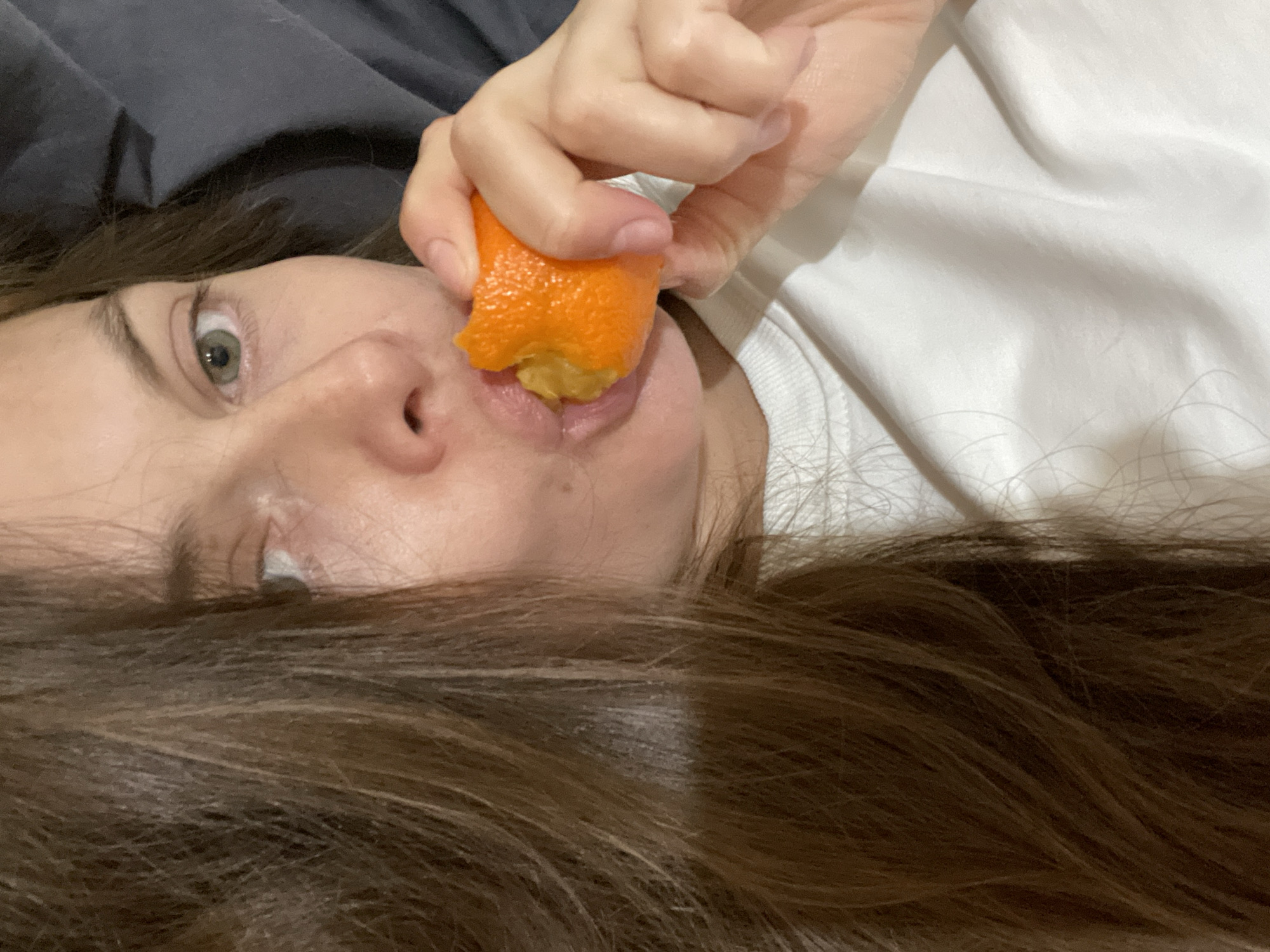 A close-up shot shows a person's face and hand as they eat a piece of orange. The person's left eye, with a greenish-gray iris, is visible, looking slightly off-camera. Their nose is also in focus, with a slight sheen on the skin. The right side of their face is mostly obscured by their hand and the orange. The person's mouth is open, with the orange partially inside. Their fingers are holding the orange, with the textured peel clearly visible. The person has long, dark brown hair that frames their face and spills across the foreground. They are wearing a white t-shirt, and a dark gray blanket or pillow is visible in the background. The scene appears to be indoors, likely a bedroom, given the soft lighting and bedding. The lighting suggests it could be daytime, but there are no strong shadows to indicate a specific time. The overall mood is casual and intimate, with the focus on the simple act of eating.