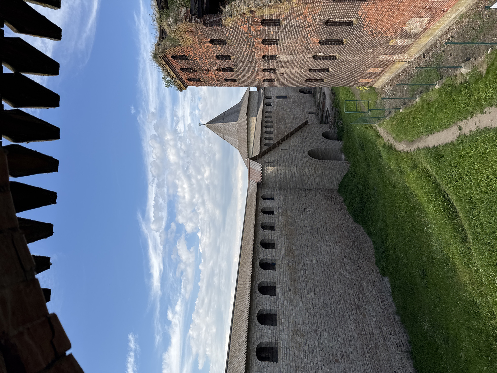The image shows a view of the Oreshek Fortress in Shlisselburg, Russia. The fortress features imposing stone walls with arched windows and a distinctive wooden-roofed tower. The top of the image is dominated by a bright blue sky with wispy white clouds, indicating clear weather and likely daytime.
In the foreground, wooden structures with a serrated edge are visible, possibly part of the fortress's defenses or observation points. The stone walls of the fortress are constructed from large, rough-hewn blocks, giving them a sturdy and ancient appearance. The wooden tower has a steeply pitched roof, capped with a small spire.
To the right of the stone walls, a green, grassy slope descends, with a path winding its way along the lower edge. Beyond this, the remains of a red brick structure are visible, overgrown with grass and showing signs of age and decay. The contrast between the weathered brick and the solid stone of the fortress suggests different periods of construction or damage.
The scene evokes a sense of historical grandeur and the enduring nature of ancient fortifications. The lighting suggests it is a sunny day, possibly mid-morning or afternoon. There is no visible text in the image. The overall impression is one of a well-preserved historical site set against a backdrop of a clear sky.
