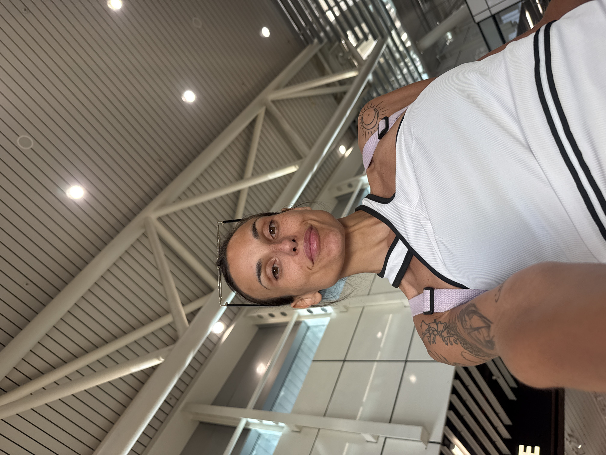 A woman with dark hair and visible freckles poses for a selfie indoors. She is wearing a white, ribbed tank top with black trim and light pink straps. She has tattoos on her left shoulder and arm, including a sun and moon design on her shoulder. She is also wearing glasses on top of her head. The background shows a modern, industrial-style ceiling with exposed beams and lighting, indicative of a large public building such as an airport terminal or convention center. The architecture features white structures and large windows. There is no specific indication in the image that places the location in Sofia, Bulgaria; this information is provided separately.