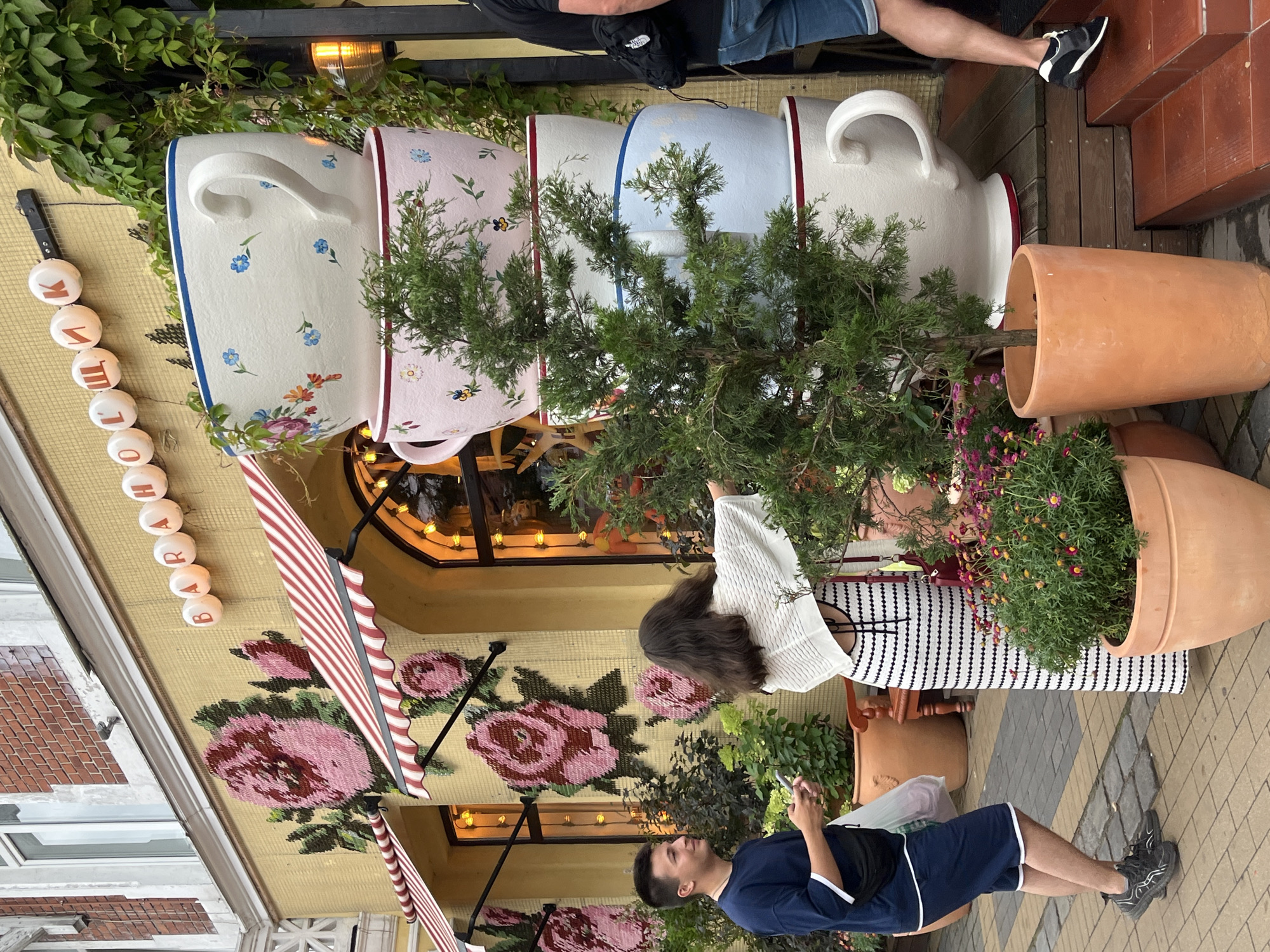 The image captures a storefront in Zelenogradsk, Russia, characterized by its yellow facade adorned with large, stylized floral mosaics, primarily featuring pink roses. Above the entrance, a row of illuminated circular letters spells out "БАРАБОШКИ" (Baraboshki). Dominating the scene are four oversized teacups stacked vertically, each decorated with floral patterns. A young man in a navy blue t-shirt and shorts stands on the paved walkway, holding a phone and a plastic bag. Beside him, a young woman in a black and white striped dress and a white shawl is reaching towards a small evergreen tree. The environment includes potted plants, a striped awning over an arched window, and wooden steps leading to the entrance. The overall impression is whimsical and inviting, likely representing a cafe or shop with a distinct theme.