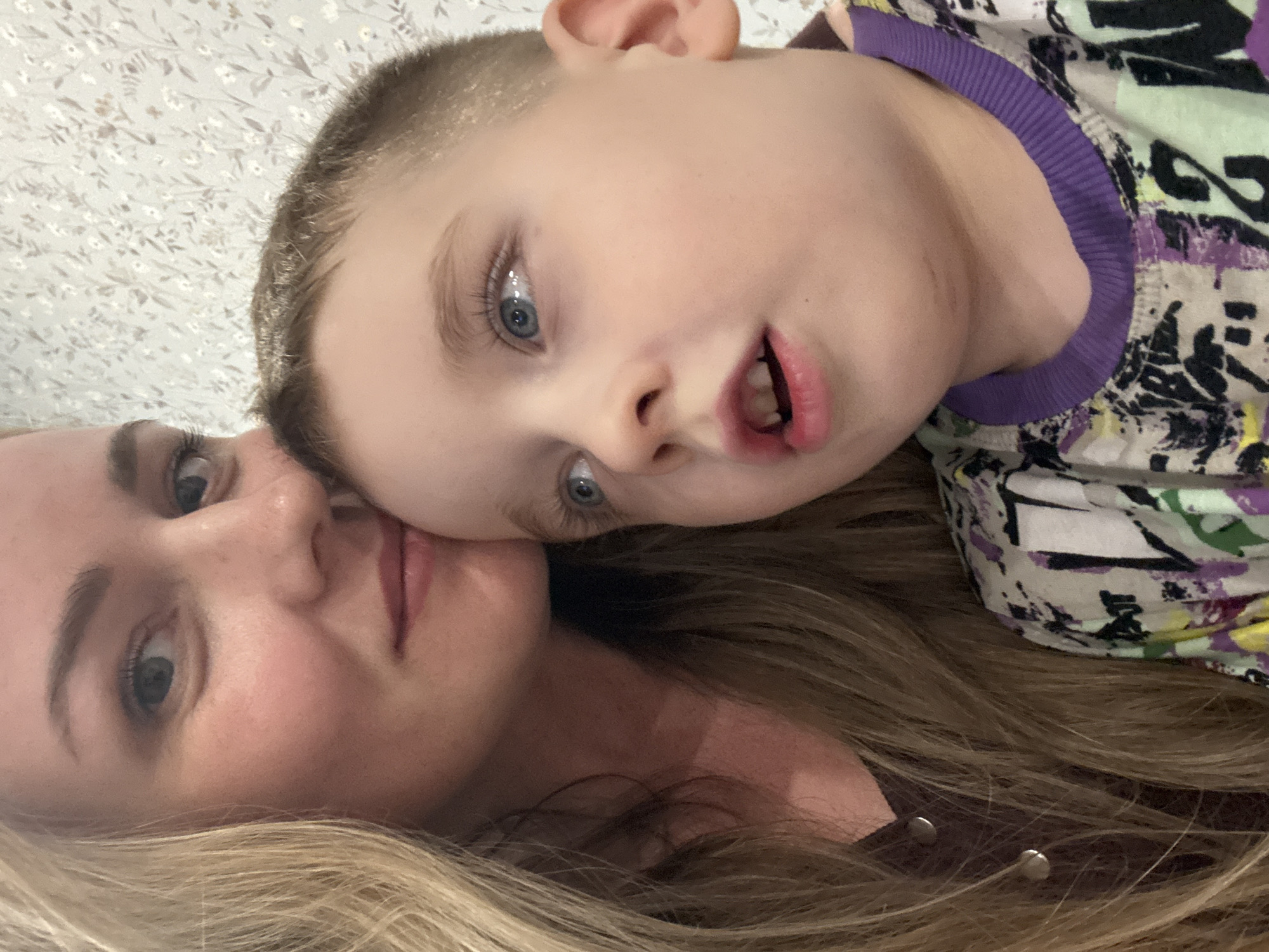 A close-up shot, likely a selfie, captures a fair-skinned woman and a young boy with blue eyes. The woman's face is in the lower portion of the frame, with long blonde hair. The boy is positioned above her, his head near her cheek. The background features a floral pattern. No information suggests a specific location in Chaikovsky, Russia.