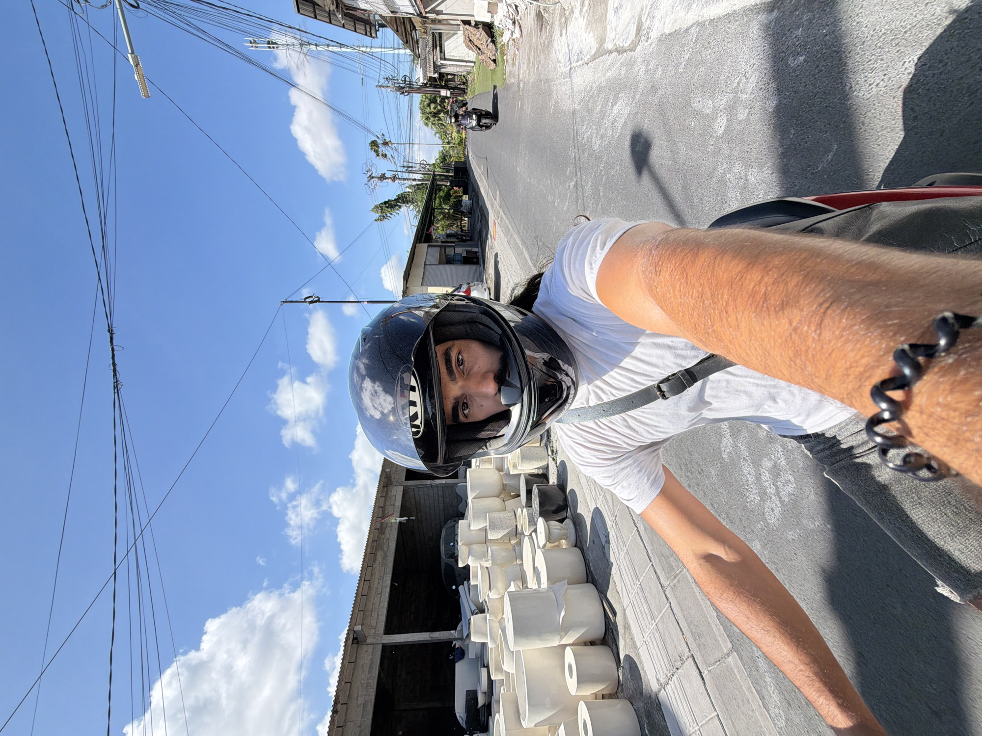 A man wearing a black helmet and white T-shirt is riding a motorcycle down a road in North Kuta, Indonesia. He is facing the camera, apparently taking a selfie. To his left is a building under construction with a stack of white objects, possibly pottery. Overhead, power lines cross the blue sky with scattered clouds. In the background, another motorcycle approaches. The road is paved.
