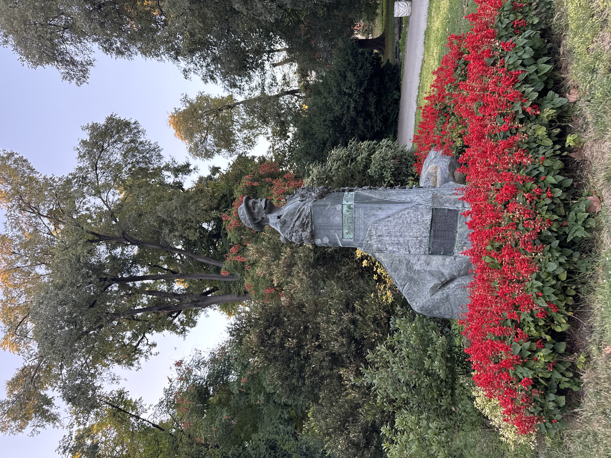 The image shows a bust of a man on a stone monument, surrounded by red flowers and lush greenery. The bust appears to be made of bronze and depicts a man with a mustache and a cap. The monument itself is a large, rough-hewn stone block with inscriptions. The setting is a park with mature trees and a paved pathway in the background. This monument is located in the Tsar Simeon Garden in Plovdiv, Bulgaria, and it commemorates the Bulgarian poet and writer Hristo Botev. The scene is captured in daylight, with a clear sky and the sun casting a soft light on the trees.