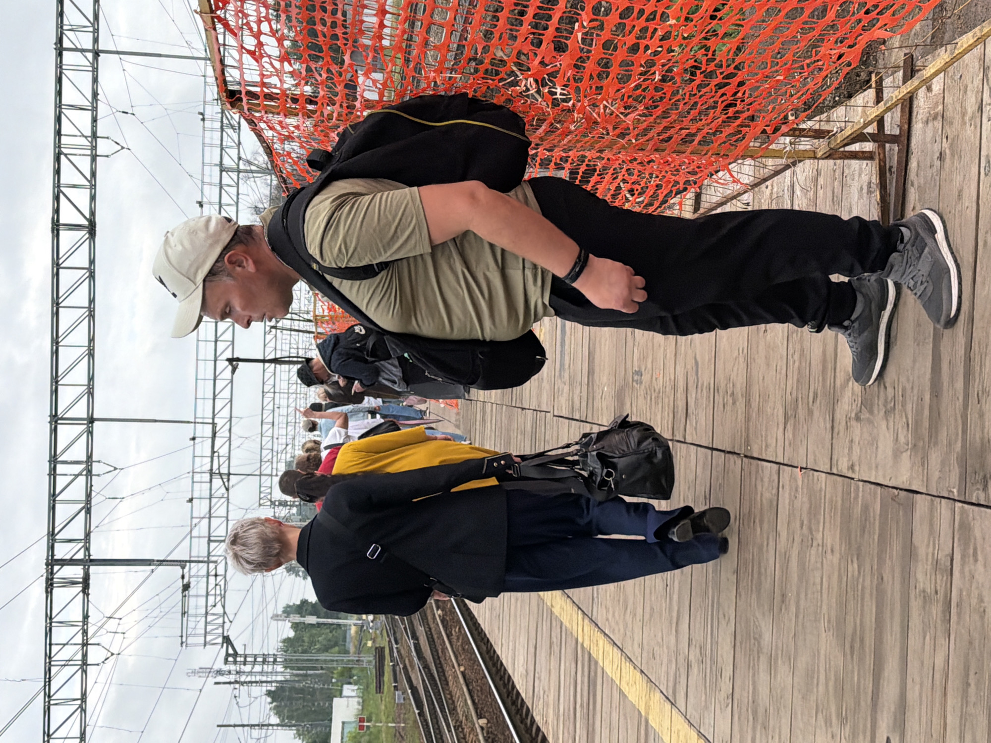 A group of people stand on a wooden train platform, waiting. In the foreground, a man wearing a beige baseball cap, a tan t-shirt, and black pants stands facing left, his back to a bright orange safety fence. He has a black backpack slung over his right shoulder. Behind him, a person wearing a dark blazer and dark pants walks away from the camera, carrying a black shoulder bag. Other individuals are visible further down the platform, some in yellow and white clothing. The platform is made of weathered wooden planks, with a yellow line marking the edge. Railroad tracks are visible to the left of the platform, and overhead wires are suspended by metal poles. The sky is overcast. The location is identified as Shcherbinka, Russia.