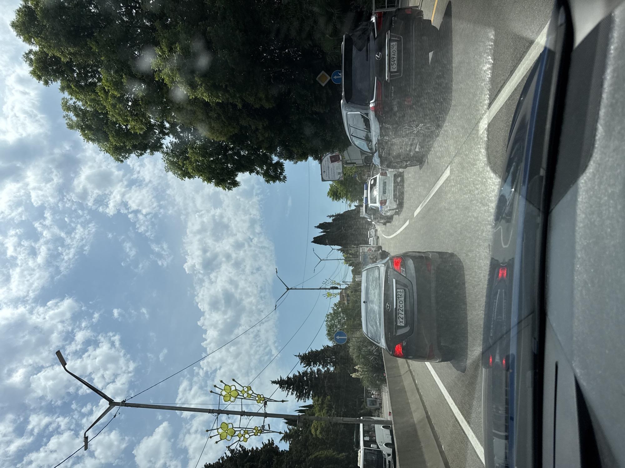 This is a photograph taken from inside a vehicle on a highway in Sochi, Russia, likely during daylight hours given the bright sky and visible clouds. The perspective is looking forward through the windshield, which has some smudges or water droplets on it.
The highway appears to be multi-lane, with a concrete barrier separating the lanes of travel and the opposing direction. A dark-colored sedan, possibly a Hyundai Solaris, is in the foreground in the left lane, with its rear brake lights illuminated. Its license plate is visible and reads "X272CO 123". To its right, in the right lane, a black Lexus LX570 SUV is visible. Its license plate reads "C549AO 46".
Further ahead, a line of traffic is visible, including a white police car with lights on top and other vehicles. On the left side of the road, there is a white bus and a concrete wall. The background features lush green trees, some of which are tall and slender like cypresses, and others with dense foliage. In the distance, there are streetlights and decorative structures with yellow, flower-like shapes suspended from poles. The sky is a bright blue with scattered white clouds. The overall scene depicts a traffic jam on a sunny day in a city with significant greenery.