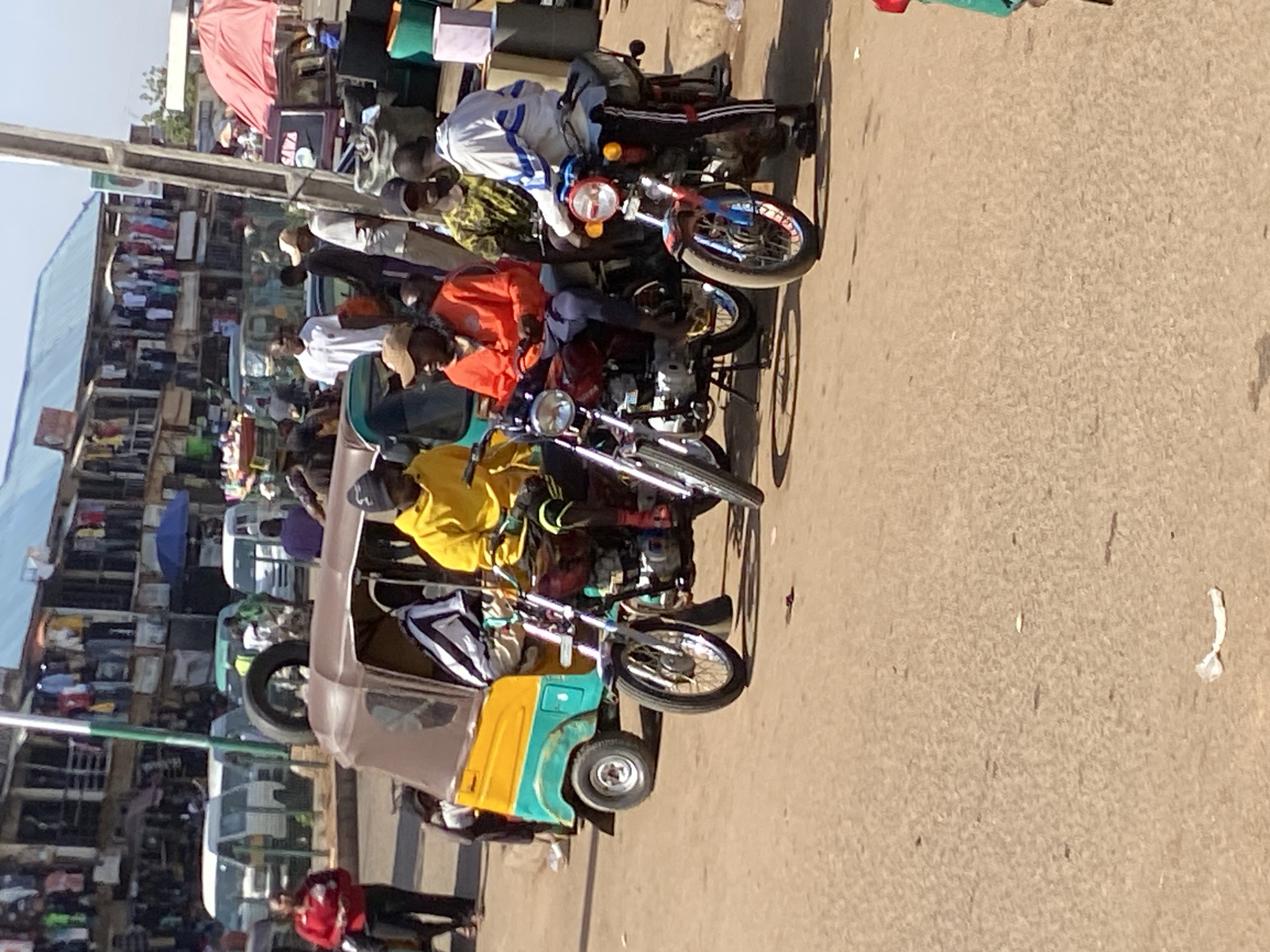 The image depicts a street scene in Bukuru, Nigeria. The street is paved and dusty. Several motorcycles and a three-wheeled auto-rickshaw are parked along the edge of the road. People are seen riding the motorcycles. A busy market is visible in the background with vendors and shoppers. Buildings line the street, and merchandise is displayed. The light suggests it is daytime.