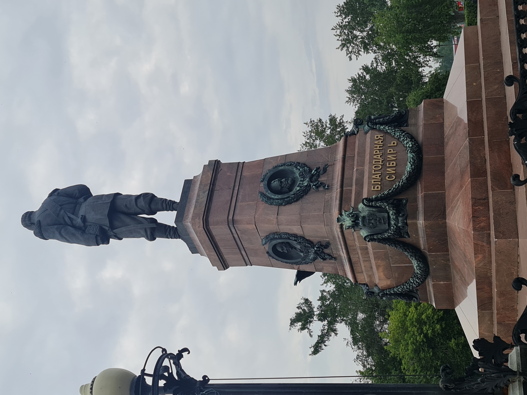 The image shows a monument with a statue of a man in military attire, viewed from behind, standing on a tall, multi-tiered pedestal. The pedestal is made of reddish-brown stone and features decorative elements, including medallions with portraits, ornate carvings, and what appears to be a gilded inscription in Cyrillic: "БЛАГОДАРНАЯ СИБИРЬ" (Gratitude to Siberia). The monument is situated outdoors, with lush green trees and an overcast sky in the background. A lamppost with a decorative base is visible to the left of the monument. This monument is the Monument to the Decembrists in Irkutsk, Russia, located in the city center.