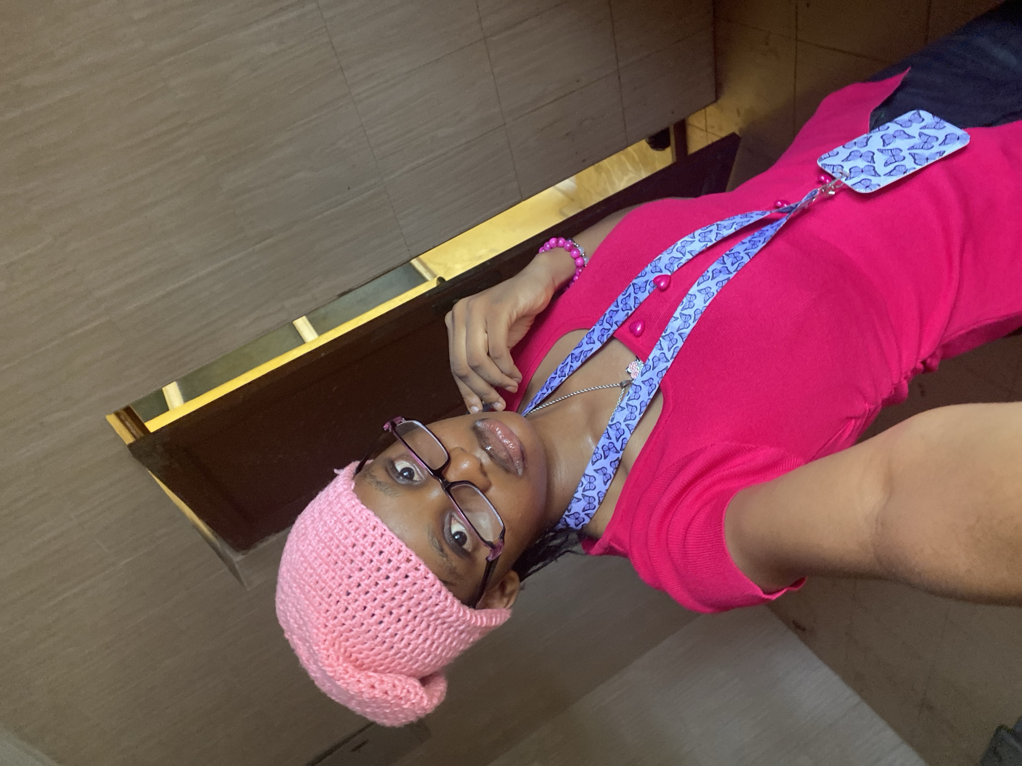 A young person wearing a pink knitted hat and glasses is taking a selfie indoors in Unknown, Unknown. They are also wearing a pink shirt, a silver necklace, and a blue lanyard with a butterfly design. The background includes a door and tiled walls.
