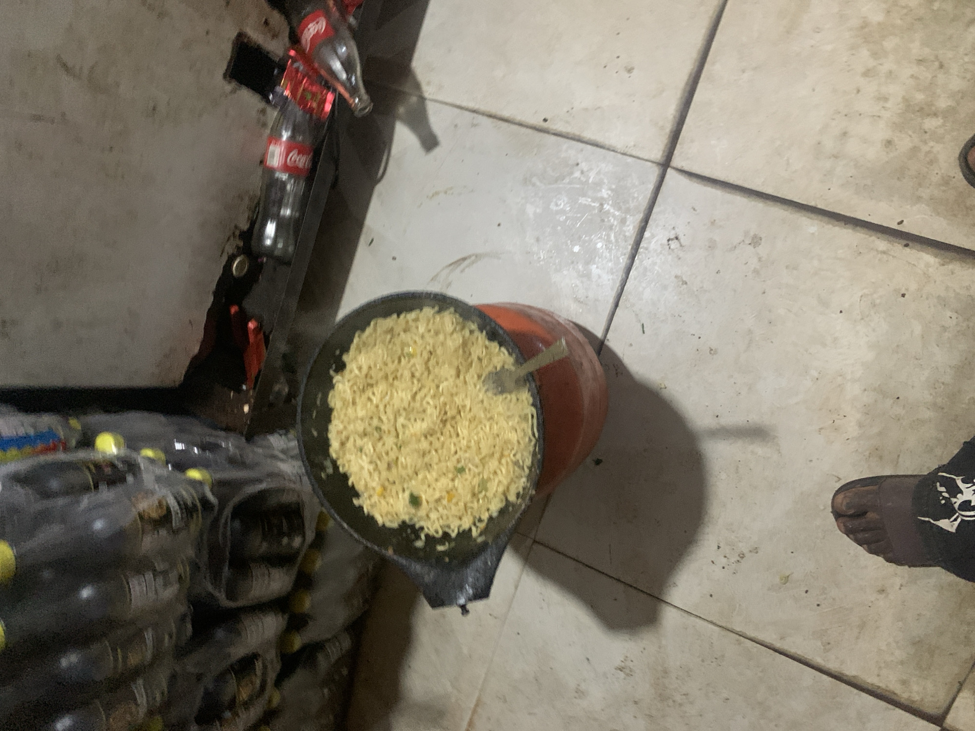 A high-angle shot captures a domestic scene in Jos, Nigeria, where cooked noodles are being prepared. In the foreground, a person's bare foot is visible, clad in a black sock with a white graphic. The individual appears to be standing on a tiled floor, which shows signs of wear and dirt.
The primary focus of the image is a pot of cooked noodles, steaming and ready to be served. The pot itself is orange and black, sitting on a stovetop or burner. The noodles are a pale yellow and appear to have some green specks mixed in, suggesting the presence of vegetables or seasonings. A fork is stuck into the noodles, indicating that they are being portioned or stirred.
Behind the pot, stacks of plastic bottles are visible, likely water or soft drinks, which are common in the Nigerian market. Further back, a shelf holds two glass bottles of Coca-Cola, one of which is lying on its side, and a red object, possibly a piece of cloth or plastic. The background is dimly lit and appears to be an indoor space, possibly a kitchen or storage area. The overall impression is that of a casual, everyday meal preparation in a home environment.