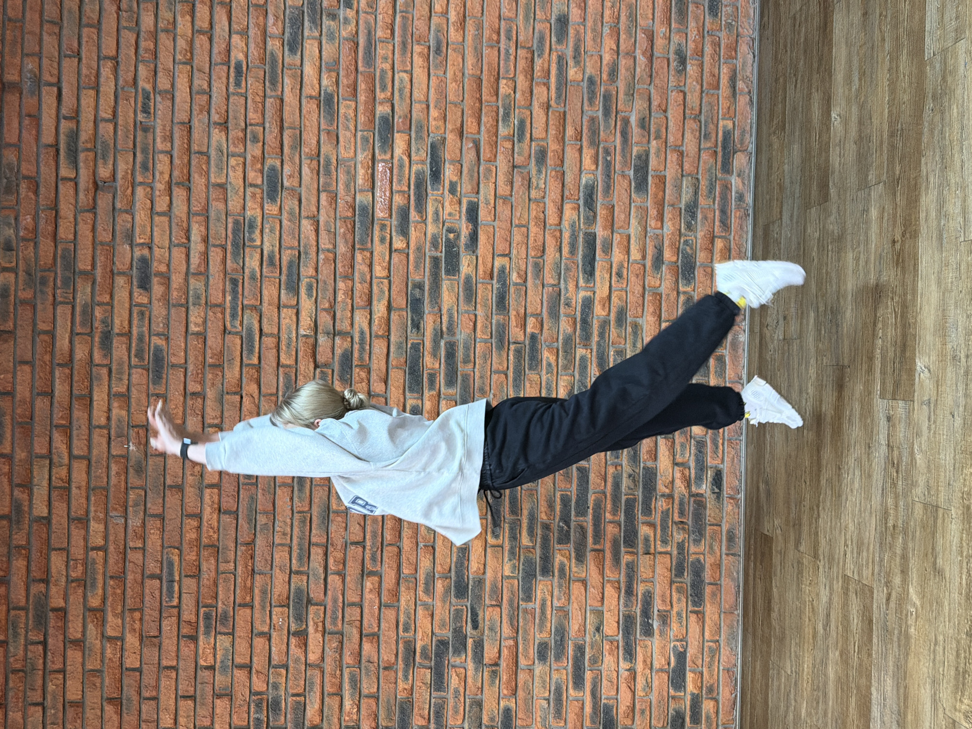 A person is depicted in a horizontal, "flying" or "floating" pose, centrally positioned in the image. The individual, appearing to be a young adult with light hair pulled back into a bun, is wearing a light grey hooded sweatshirt, dark athletic pants, and white athletic shoes with subtle yellow accents. A dark fitness tracker or smartwatch is visible on their left wrist.
The background is divided vertically into two distinct textures. The left side features a brick-patterned surface composed of reddish-brown bricks with irregular dark grey or black patches, laid out in vertical rows. The right side shows a wooden surface with planks running vertically, separated from the brick area by a thin dark border.
The activity suggests a staged photograph aiming for an illusion of levitation or flight, with the person's body elongated, arms extended forward with hands slightly open, and legs stretched backward and slightly splayed. The dynamic pose conveys a sense of weightlessness and motion.
The setting appears to be indoors, under even lighting, with no visible external weather or time-of-day indicators. While the image's orientation suggests the person is flying against a vertical brick wall with a wooden floor to the side, it could also represent a person lying on a textured ground photographed from above and then rotated for artistic effect. The provided location of Chaikovsky, Russia, is not visually evident within the image itself. There is an indistinct logo on the front of the sweatshirt, but no other readable text is visible.