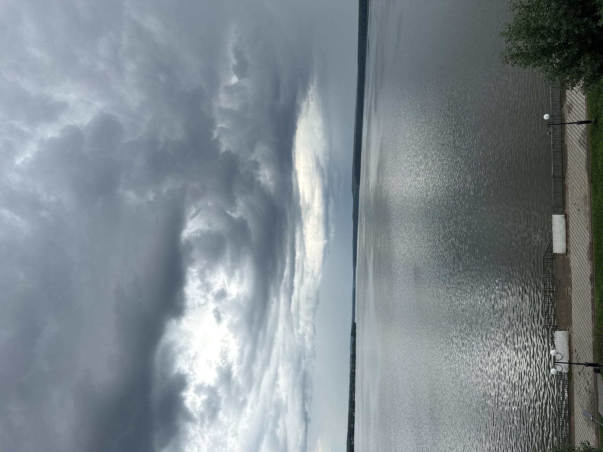 The image captures a wide shot of a body of water with a cloudy sky above. The left side of the frame is dominated by dark, dramatic clouds, suggesting an impending storm or overcast weather. Sunlight breaks through in patches, illuminating the cloud formations. A faint speck, possibly a bird, is visible in the sky.
The right side of the image shows a vast expanse of water, likely a river or lake, with a shimmering surface due to light reflecting off ripples. The water appears calm but textured. Along the edge of the water, there's a paved walkway with a decorative pattern of light and dark tiles. A black metal railing separates the walkway from the water. A black lamppost with two globe-shaped lights stands on the walkway, casting faint shadows. To the far right, the edge of a leafy green tree is visible. In the distance, across the water, a dark line of trees or buildings forms the horizon, suggesting a shoreline or the opposite bank. The overall mood is serene yet somewhat somber due to the heavy clouds. The time of day is difficult to determine precisely but appears to be daytime, possibly late afternoon or early evening, given the quality of light and the cloudy conditions. No visible text is present in the image.