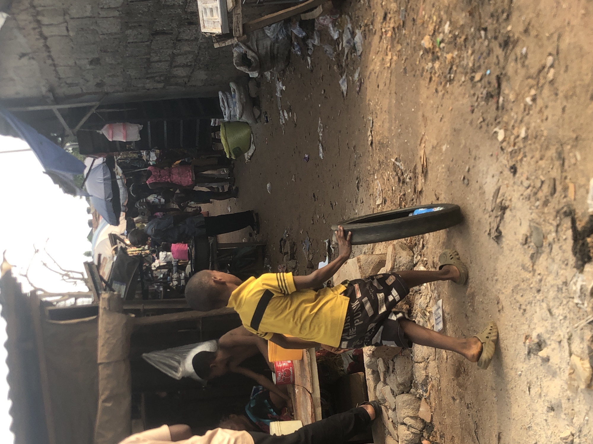 This image captures a vibrant, busy outdoor scene, likely a market or a densely populated street in Onitsha, Nigeria, during the daytime. The ground is unpaved, consisting of packed dirt, scattered rocks, and various debris and litter.
The central subject is a young boy, positioned towards the right side of the frame. He is wearing a bright yellow short-sleeved polo shirt with black stripes on the shoulders and sleeves, and patterned shorts. Text visible on his shorts includes "PLAN-C" and "THE PUNK." He wears light-colored slip-on sandals. His right hand rests on a dark tire lying on the ground. To his left and slightly behind him, another boy, shirtless, is visible, looking downwards towards an obscured area.
In the background, a bustling environment unfolds. Numerous people are present, some partially obscured by makeshift market stalls and structures. These structures are constructed from materials like concrete blocks, wood, and corrugated metal sheets, some covered by fabric or tarps. Various goods, possibly clothing or household items, are displayed for sale. Several umbrellas, including an open blue one and some folded ones, are scattered throughout the scene, suggesting a need for shade or shelter. A yellow bucket and a red plastic container are also visible on the ground near the boys. The scene portrays daily life and commerce in a dense, urban setting.