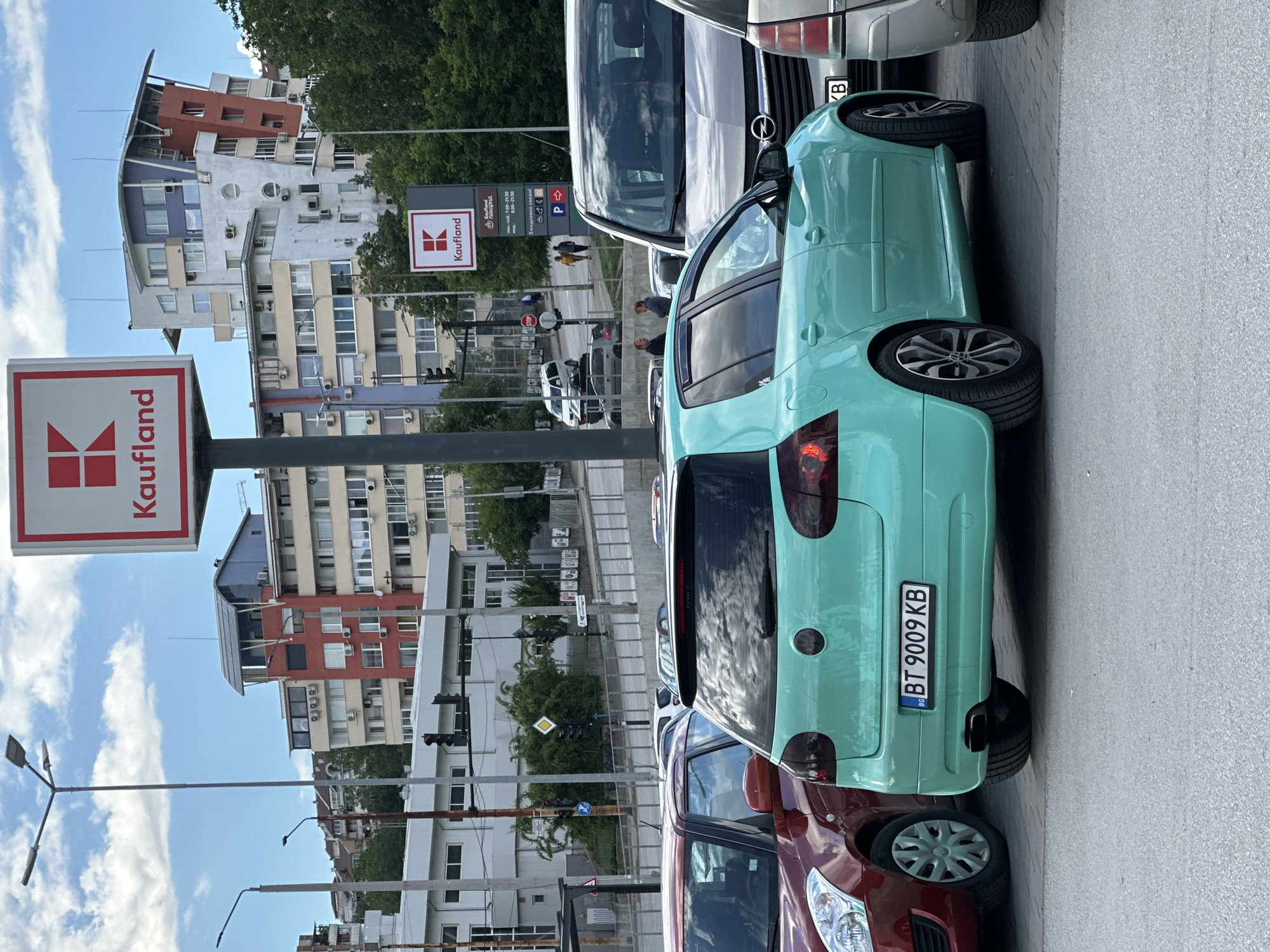 A teal Volkswagen Golf hatchback with license plate BT 9009 KB is parked in what appears to be a parking lot in Veliko Tarnovo, Bulgaria. The car is facing away from the camera, and its rear bumper, taillights, and alloy wheels are visible. To the left of the Golf, a red Suzuki Swift is parked. In the background, several modern buildings with mixed architectural styles can be seen, some featuring colorful facades and balconies. A large red and white sign for "Kaufland" is prominently displayed on a pole in the foreground. Additional traffic lights and street signs are visible in the midground, suggesting an urban or commercial area. The sky is partly cloudy.