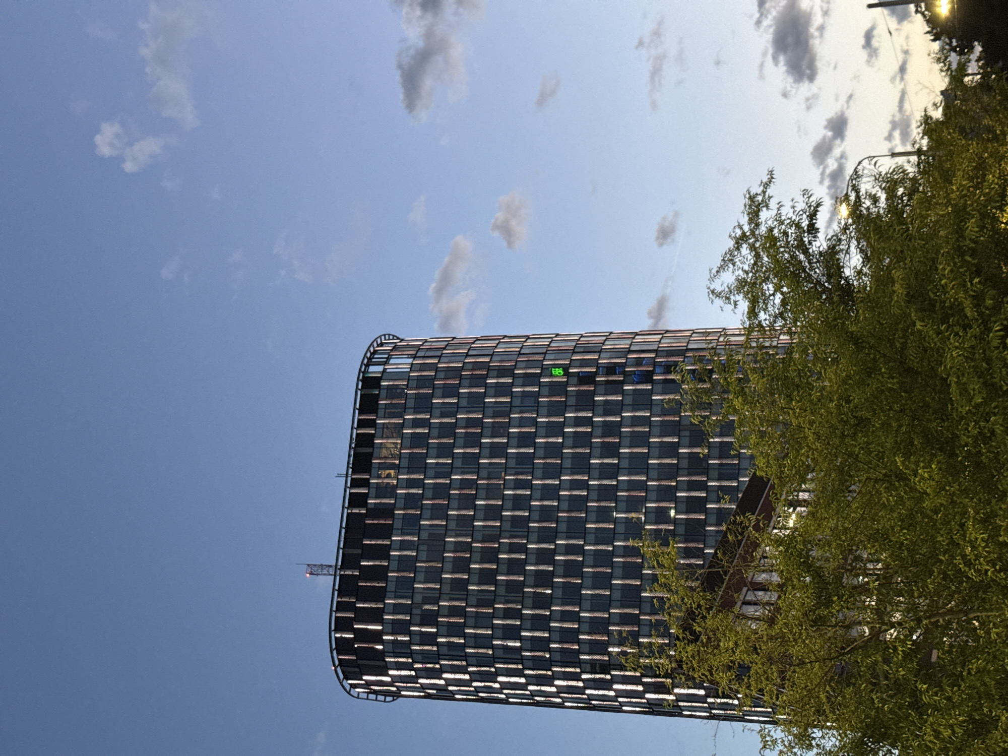 In Bratislava, Slovakia, a tall, cylindrical skyscraper with a reflective facade stands against a twilight sky. The building's exterior is composed of numerous vertical panels, creating a pattern that reflects the ambient light. A few scattered clouds drift across the sky, illuminated by the fading sunlight. In the foreground, the dense green foliage of trees obscures the lower portion of the skyscraper, their leaves rustling gently. A streetlamp is visible through the trees, casting a warm glow. The overall scene depicts an urban environment transitioning from day to night.