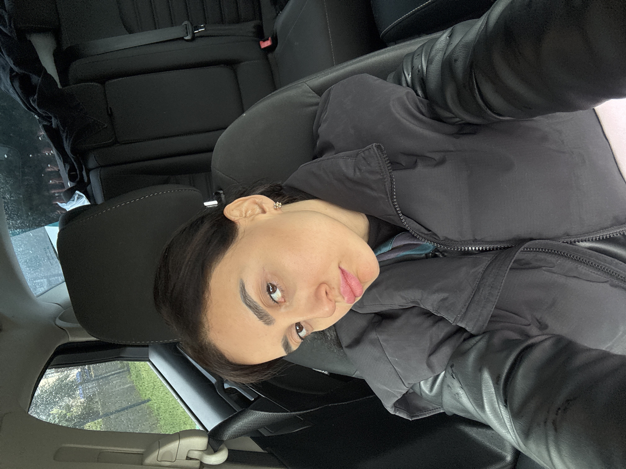 A woman with fair skin and dark hair is positioned in the driver's seat of a car. She is wearing a black jacket with what appears to be leather sleeves and is looking towards the camera with a slight pout. The interior of the car is visible, including the black seats and headrests, as well as a rear seat with a seatbelt. Through the window, there's a view of what appears to be a rainy or wet outdoor environment with some green foliage. There is no evidence of location in Bolokiani, Georgia.