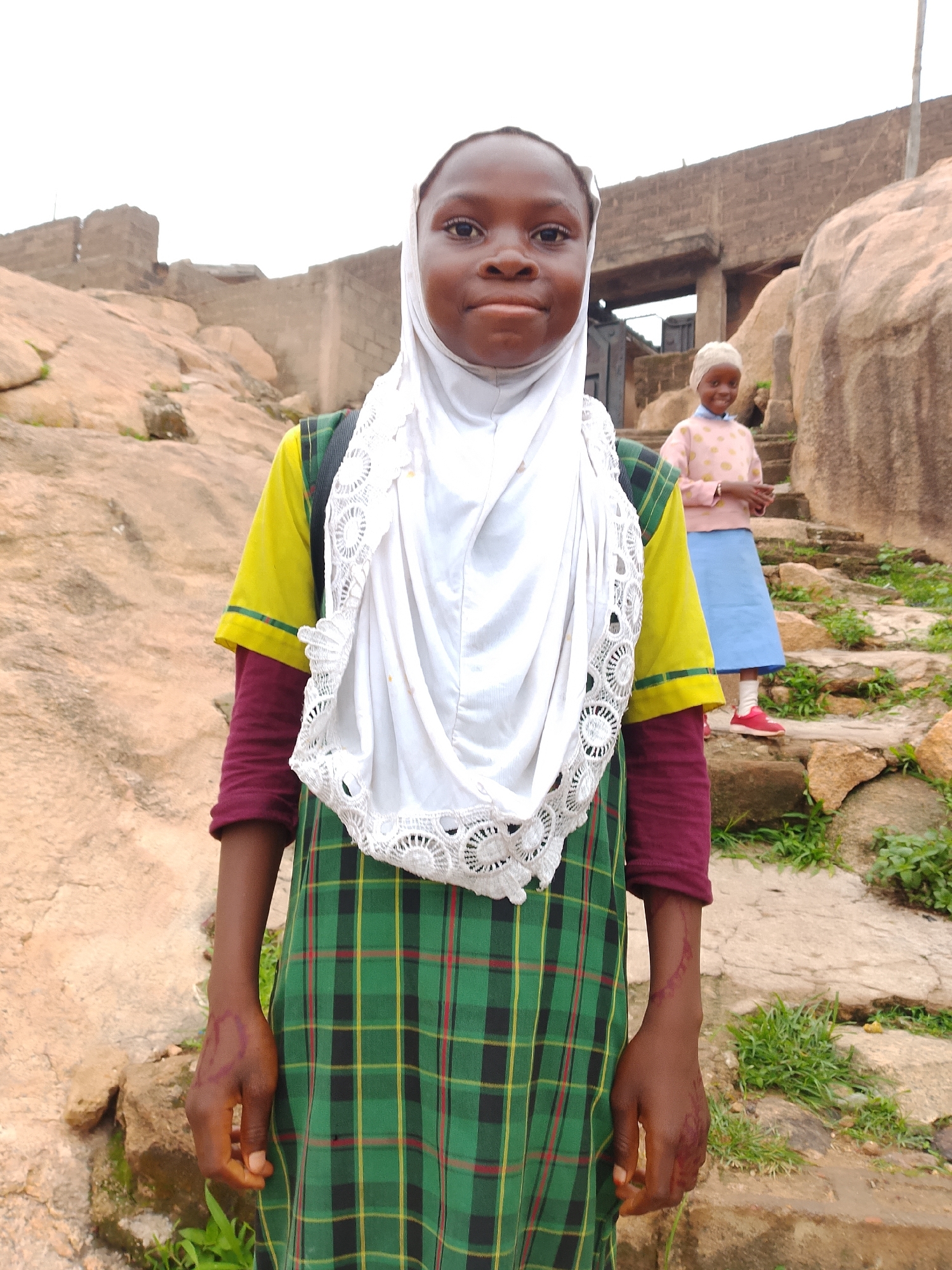 A medium shot shows two young girls standing on rocky terrain with buildings and rock formations in the background. The girl in the foreground is wearing a white hijab, a yellow and maroon long-sleeved shirt with a green and black plaid dress. She is looking directly at the camera with a neutral expression. Her hands are visible and appear to have some red markings on them. To her right and slightly behind her, another young girl is visible. She is wearing a light pink shirt with yellow polka dots and a light blue skirt. She is looking down and to her right with a slight smile. The environment appears to be an outdoor setting with rocky ground, some greenery, and structures that suggest an urban or semi-urban location. The sky is overcast.
