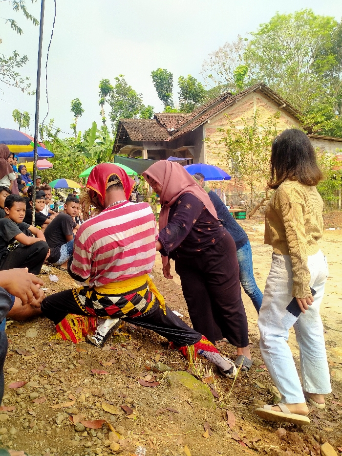 The image depicts an outdoor scene in the city of Lempong, Indonesia. The foreground consists of uneven, reddish-brown dirt ground scattered with dry leaves and small rocks. In the central midground, a person, viewed from behind, is in a low, squatting posture with legs extended. This individual wears a red and white horizontally striped long-sleeved shirt, black pants, and a yellow and black patterned fabric tied around their waist. Red and yellow fringed adornments are visible near their ankles, and a red cloth covers their head. To the immediate right, a woman wearing a dark, patterned long-sleeved top and a pink hijab leans forward towards the central figure. Further to the right and slightly behind, another woman, viewed from behind, stands with shoulder-length dark hair, wearing a light brown long-sleeved top and light-colored jeans, holding a dark smartphone in her left hand. A man in a blue shirt and blue jeans is partially visible behind the woman in the hijab. In the left background, a group of individuals, predominantly young males, are seated on the ground under various colorful umbrellas (blue, green, and multi-colored), observing the scene. The background features lush green foliage, tall trees, and a single-story house with a red tiled roof and light-colored brick walls. A slender vertical pole or wire extends upwards on the far left of the frame.