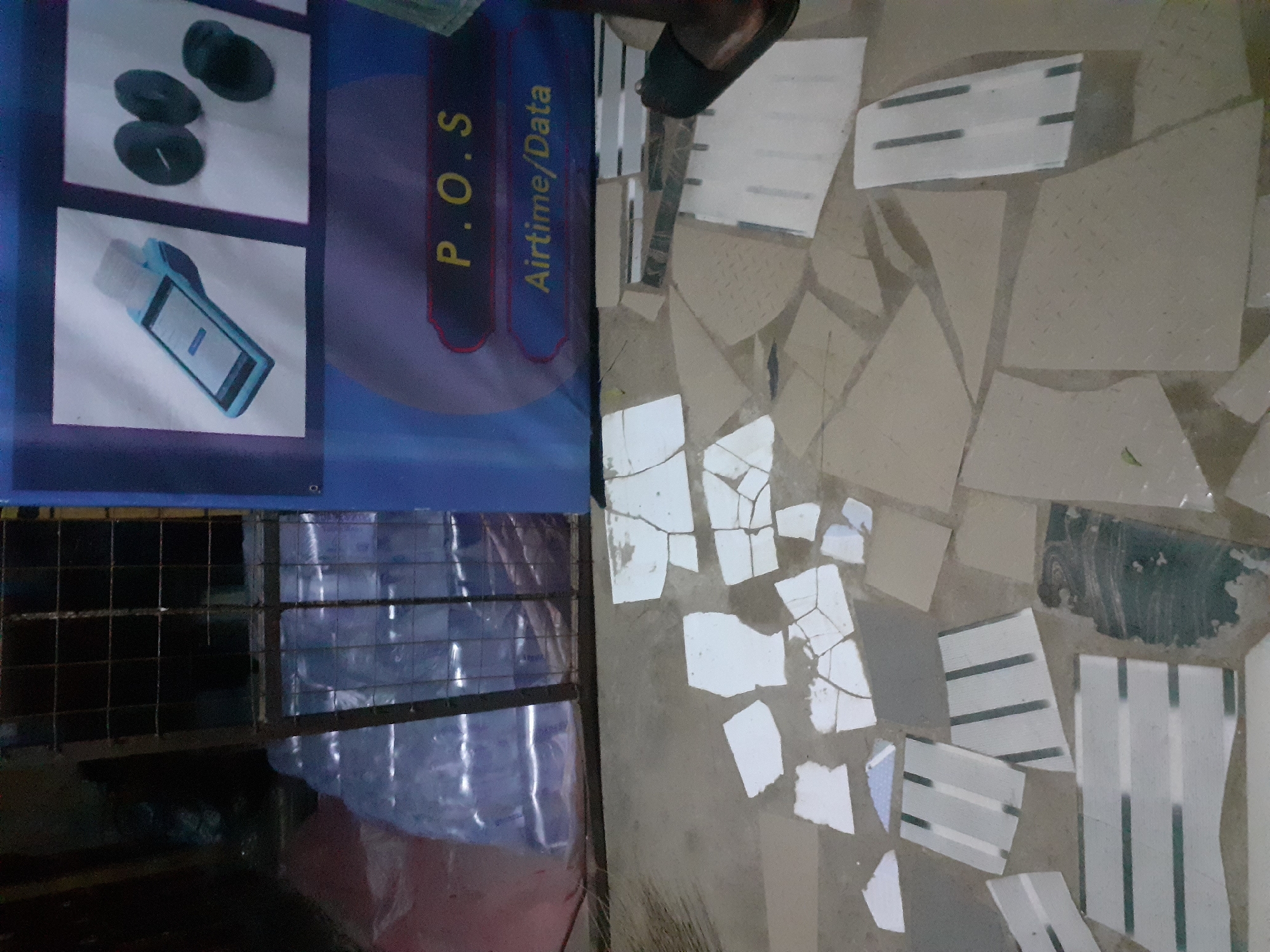 The image depicts a street-level scene in Jalingo, Nigeria. In the background, a blue banner advertises "P.O.S Airtime/Data" and displays images of a point-of-sale device and small black circular objects. Adjacent to the banner is a cage-like structure with a metal grate, behind which stacks of bottled water are visible. The foreground shows a broken tiled floor with numerous scattered pieces of white and grey tiles, some of which appear to be decorated with dark stripes. A person's foot wearing a black sandal is visible on the right side of the image. The overall impression is of an outdoor commercial area with signs of damage or disrepair in the flooring.