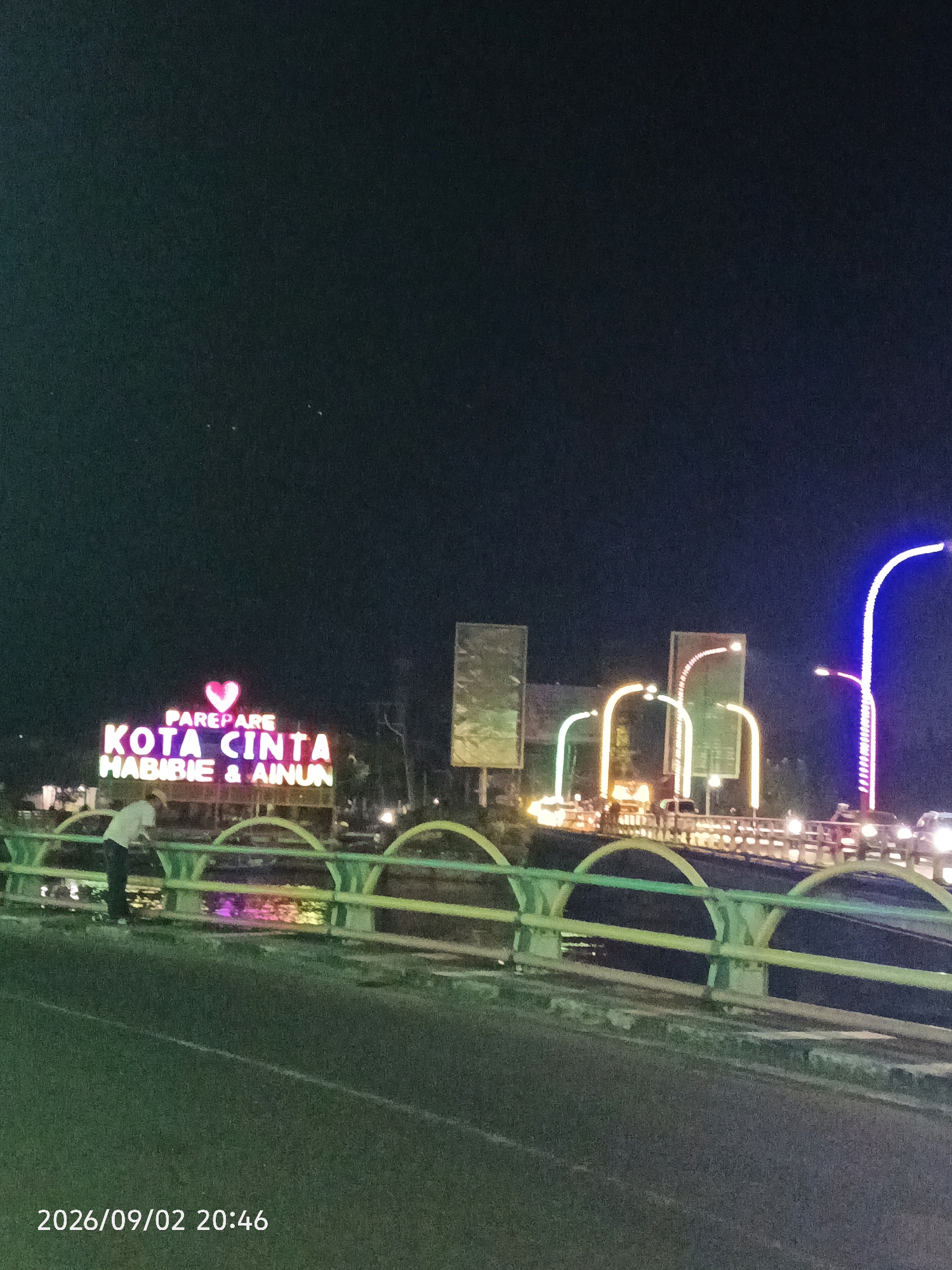 A nighttime, low-angle shot captures a illuminated bridge decorated with colorful lights in Pare-Pare, Indonesia. Dominating the center of the frame is a neon sign that reads "PAREPARE KOTA CINTA HABIBIE & AINUN," featuring a pink heart symbol above the text. The sign is lit with a combination of pink, purple, and yellow hues. Below the sign, a person in a white shirt and dark pants leans over the bridge railing, their back to the camera. The bridge itself is bordered by a light green railing with yellow accents. In the background, other illuminated structures and streetlights, including a tall, brightly lit purple lamppost on the right, line the roadway. The scene is set against a dark night sky. The date and time stamp "2026/09/02 20:46" is visible in the lower left corner.
