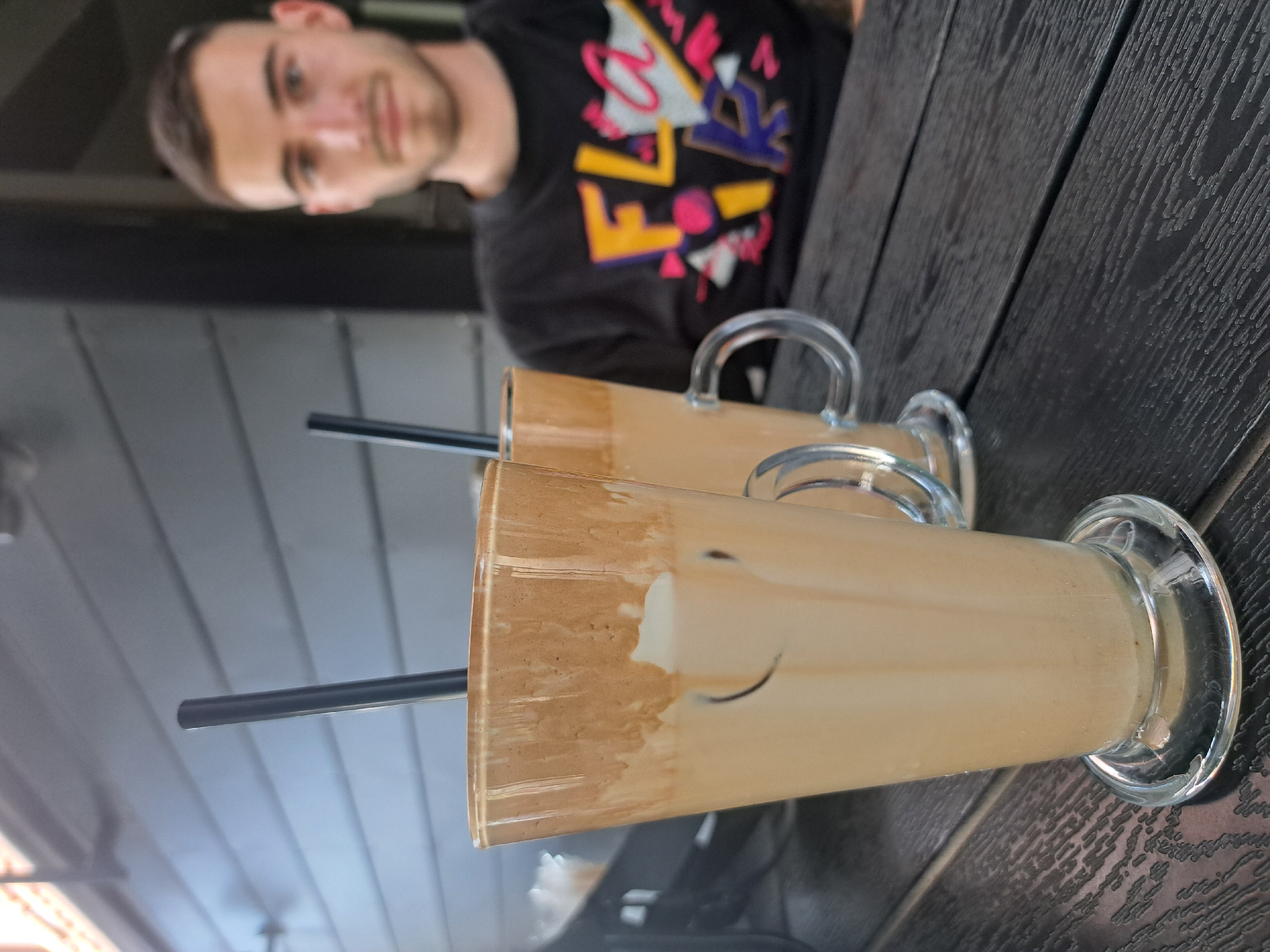 A person is seen from the shoulders up, looking towards the camera with a neutral expression. They have short dark hair and are wearing a black t-shirt with a colorful, abstract graphic design on it. The text "FLYER" is visible in large yellow lettering on the shirt.
In the foreground, two tall glasses of a creamy, light brown beverage are prominently featured. Each glass has a clear handle and a dark straw. The beverage appears to be a cold coffee drink, possibly dalgona coffee, with a frothy top. The glasses are placed on a dark, textured surface that resembles a wooden table.
The background is out of focus, but suggests an outdoor setting with some overhead structures that appear to be slatted or paneled, possibly a patio or a covered seating area. The lighting suggests it could be daytime. The overall mood is casual and relaxed.