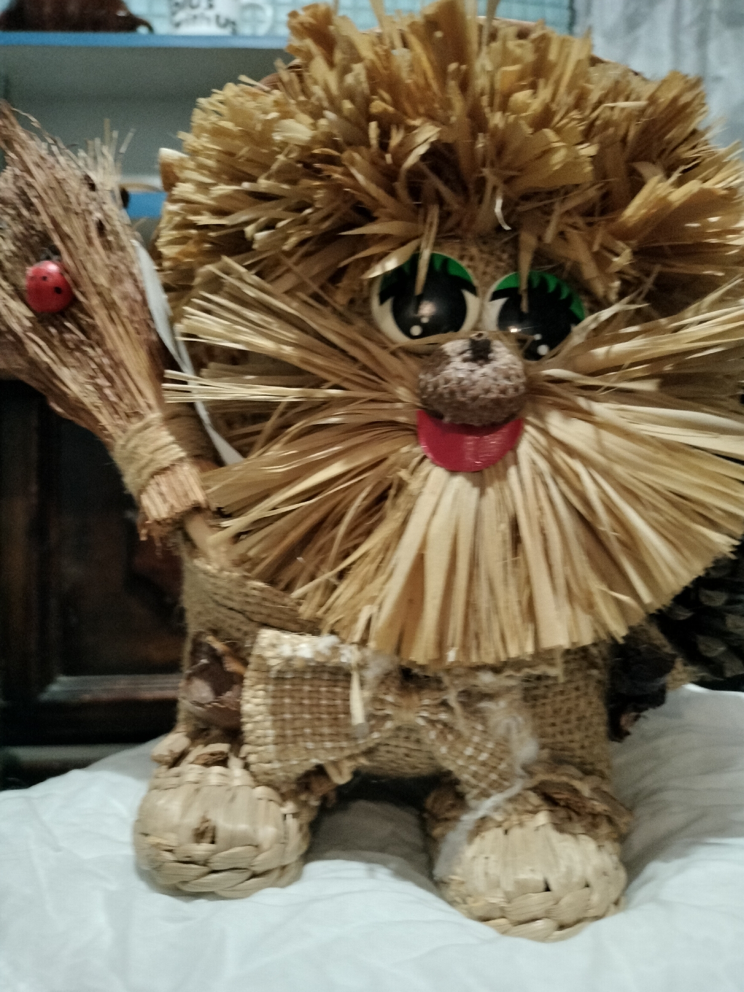 A handmade straw figurine of a lion-like creature is displayed indoors on a white cloth surface. The figurine has a mane made of straw, large green eyes with black pupils, a small brown nose, and a red mouth. It is holding a miniature broom with a red ladybug attached to it in its left paw. The body of the figurine is covered in burlap material and straw. The background appears to be a shelf with various items, some of which are out of focus. The figurine is positioned centrally in the frame, facing forward. No people or specific location details within Lykhove, Ukraine are visible.