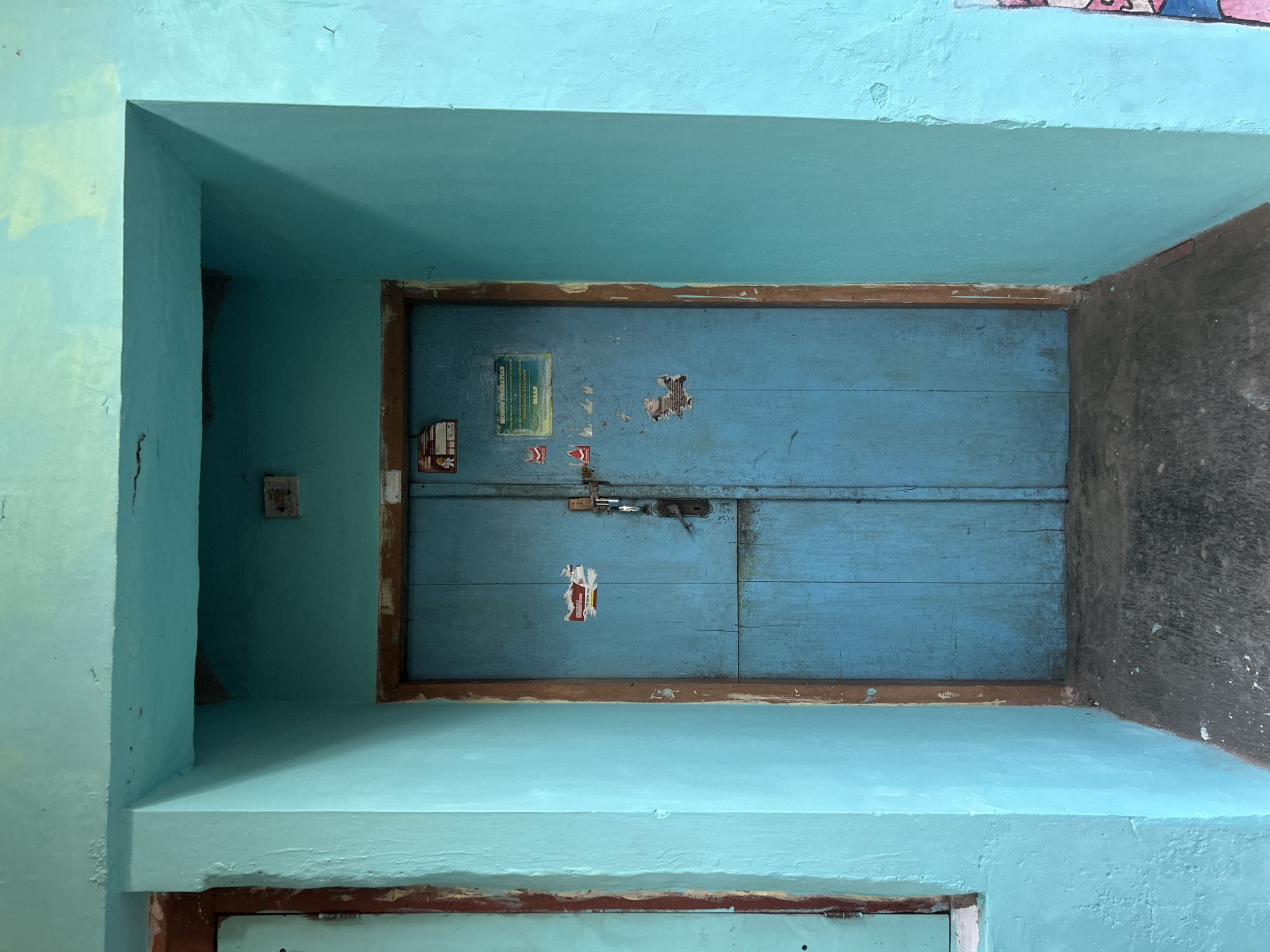 The image shows a closed, blue wooden double door set within a teal-colored alcove. The door is weathered and has several stickers and what appears to be a padlock and latch. The surrounding walls are painted teal, with some areas showing signs of wear and tear. The ground in front of the door is a dark, rough surface. The scene appears to be outdoors, possibly in an alleyway or entryway in Yogyakarta, Indonesia. There are no people or animals visible in the image.
