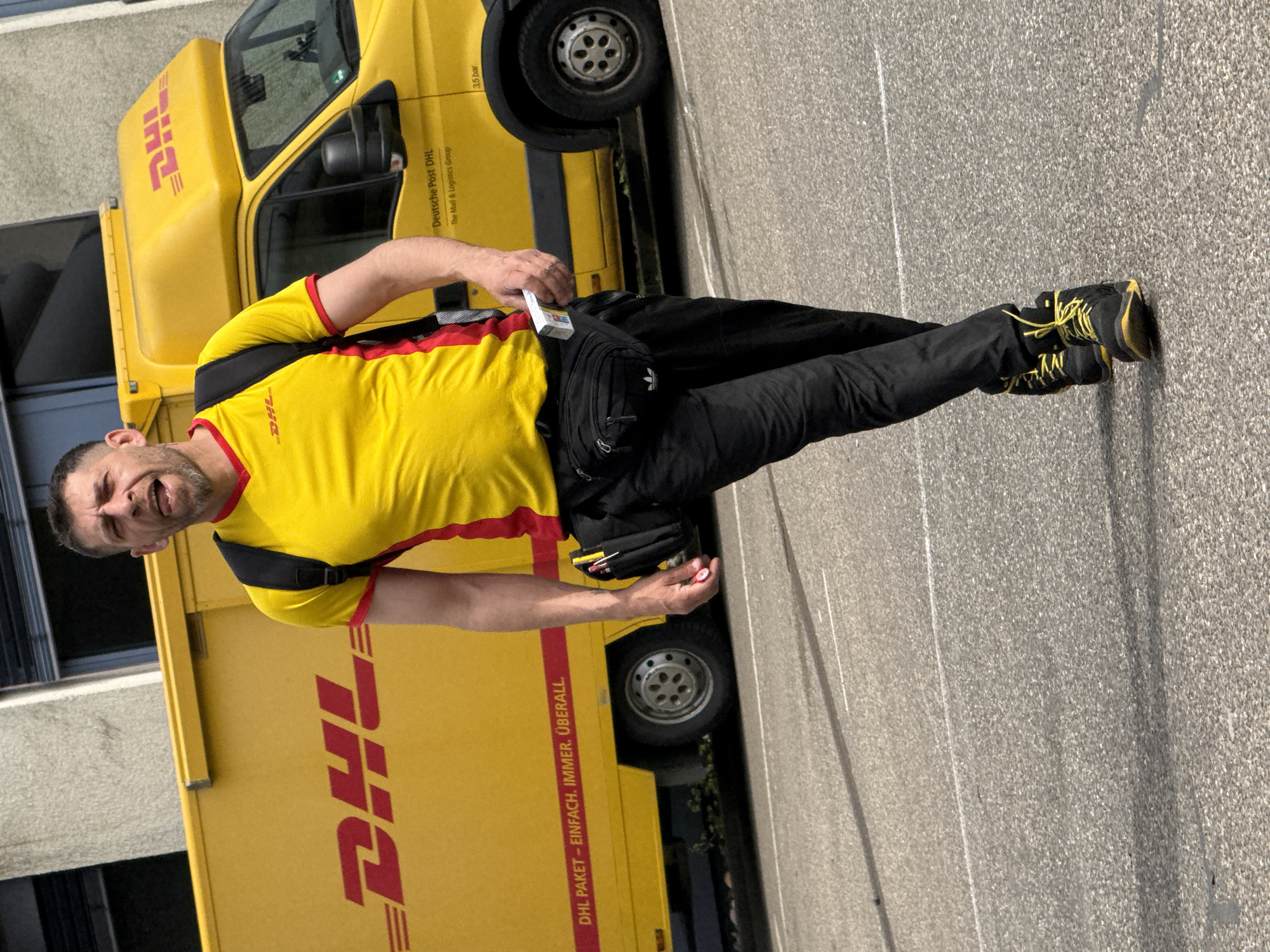A male DHL delivery driver is captured standing next to his yellow DHL delivery van in an outdoor setting, likely a paved street or loading area in Bietigheim-Bissingen, Germany. The image is taken during bright daylight hours.
The man, appearing to be in his 30s or 40s, has a short beard and mustache. He is wearing a yellow short-sleeved uniform shirt with red trim, featuring the "DHL" logo prominently. He also wears black trousers and black sneakers with distinctive bright yellow laces. A black fanny pack with an "A" logo (likely Adidas) is slung across his chest/shoulder, and another black bag is secured at his waist. He looks directly at the camera with an animated expression, his mouth open, possibly mid-speech or making a playful face. In his right hand, he holds a white packet of cigarettes.
The bright yellow delivery van behind him is clearly branded with the "DHL" logo. Text on the vehicle includes "DHL PAKET - EINFACH. IMMER. ÜBERALL." (DHL PARCEL - SIMPLE. ALWAYS. EVERYWHERE.) along its side, and "Deutsche Post DHL" with "The Mail & Logistics Group" on the cab door. A "3.5 bar" marking is visible near a tire. The scene portrays a casual moment during the driver's work, possibly a brief pause or interaction.
