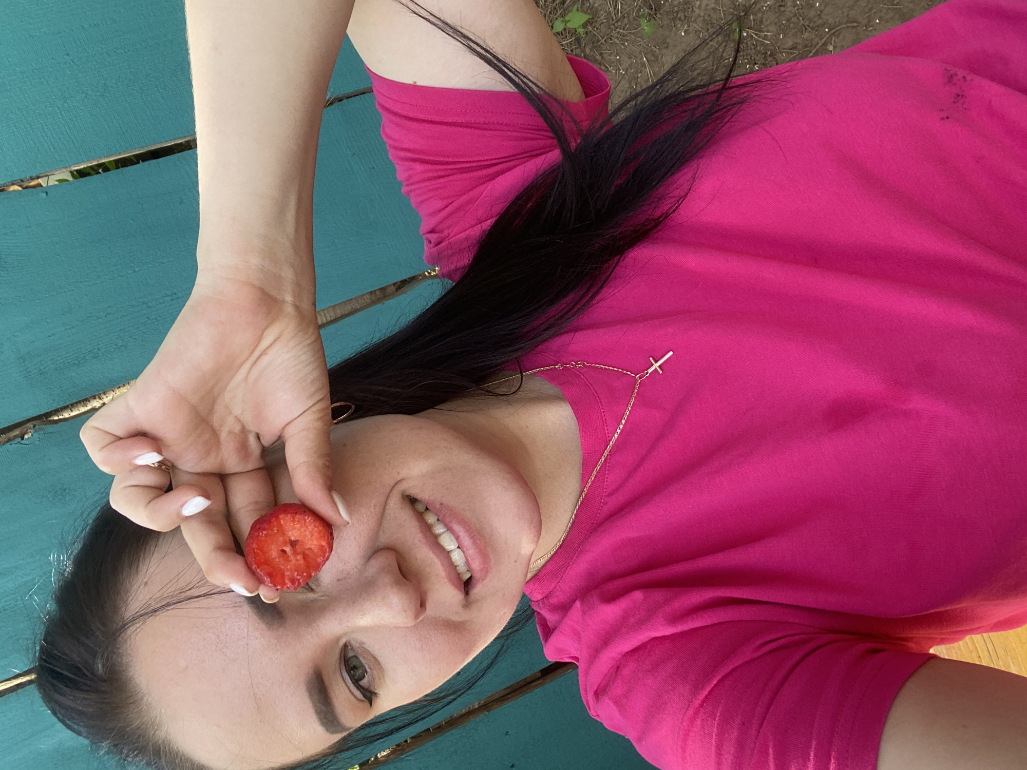 A woman with long, dark hair and a pink t-shirt is lying on a turquoise wooden surface, possibly a picnic table. She holds a cut strawberry in front of her right eye with her left hand, displaying white nail polish on her fingernails. She smiles slightly, showing her teeth. A thin gold necklace with a cross pendant is visible around her neck. The background appears to be a mix of wood and possibly some ground with sparse vegetation, suggesting an outdoor setting. The image does not provide any information about the location in the city of Chaikovsky, Russia.