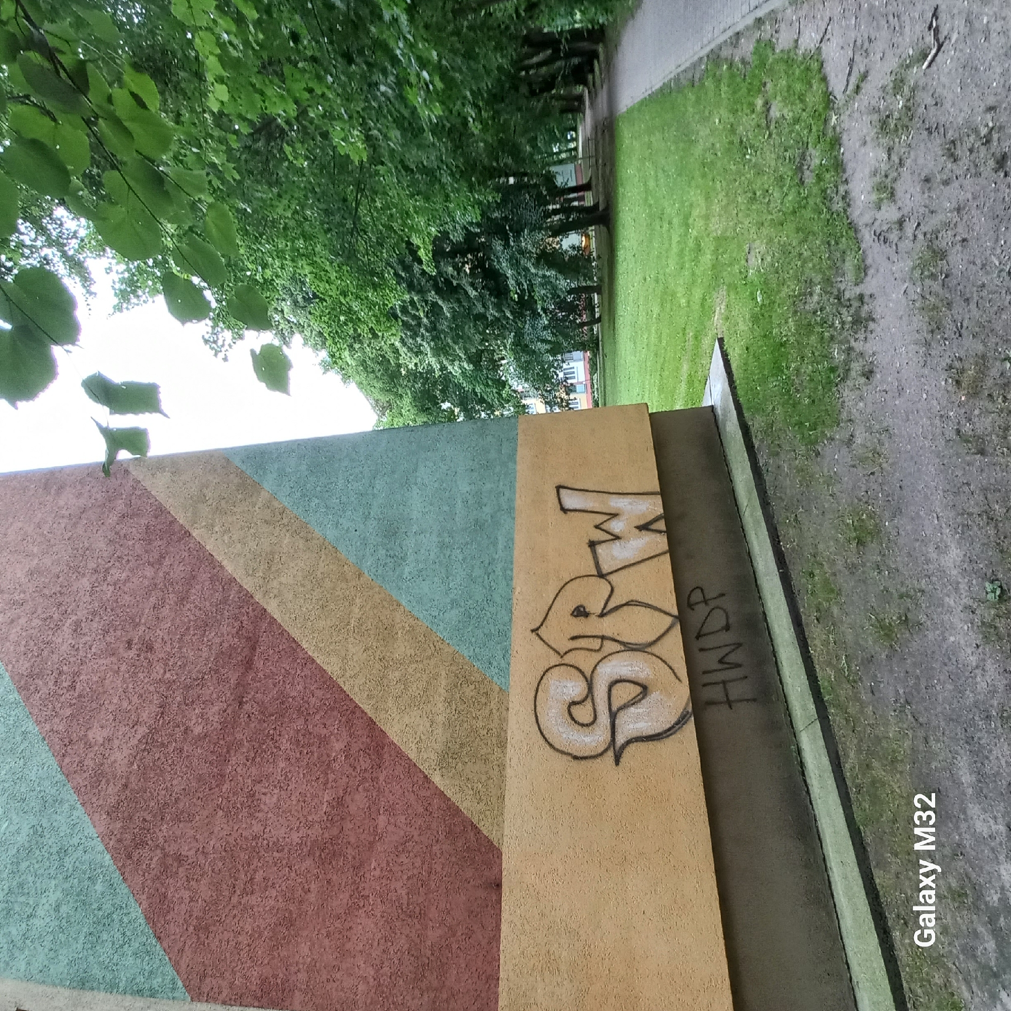 The image shows a section of a building with abstract geometric patterns in shades of pink, yellow, and teal. On the lower, tan-colored section of the building, there is graffiti. The graffiti on the top part of the tan section appears to be in white with black outlines and reads "SPRW". Below this, there is more graffiti in black, reading "HWDP". The building is situated next to a grassy area with trees and a paved walkway. In the background, other buildings can be seen, along with more trees. The scene is outdoors, likely in a residential area of Olsztyn, Poland, given the metadata. The lighting suggests it is daytime, possibly overcast, with visible green foliage from deciduous trees.