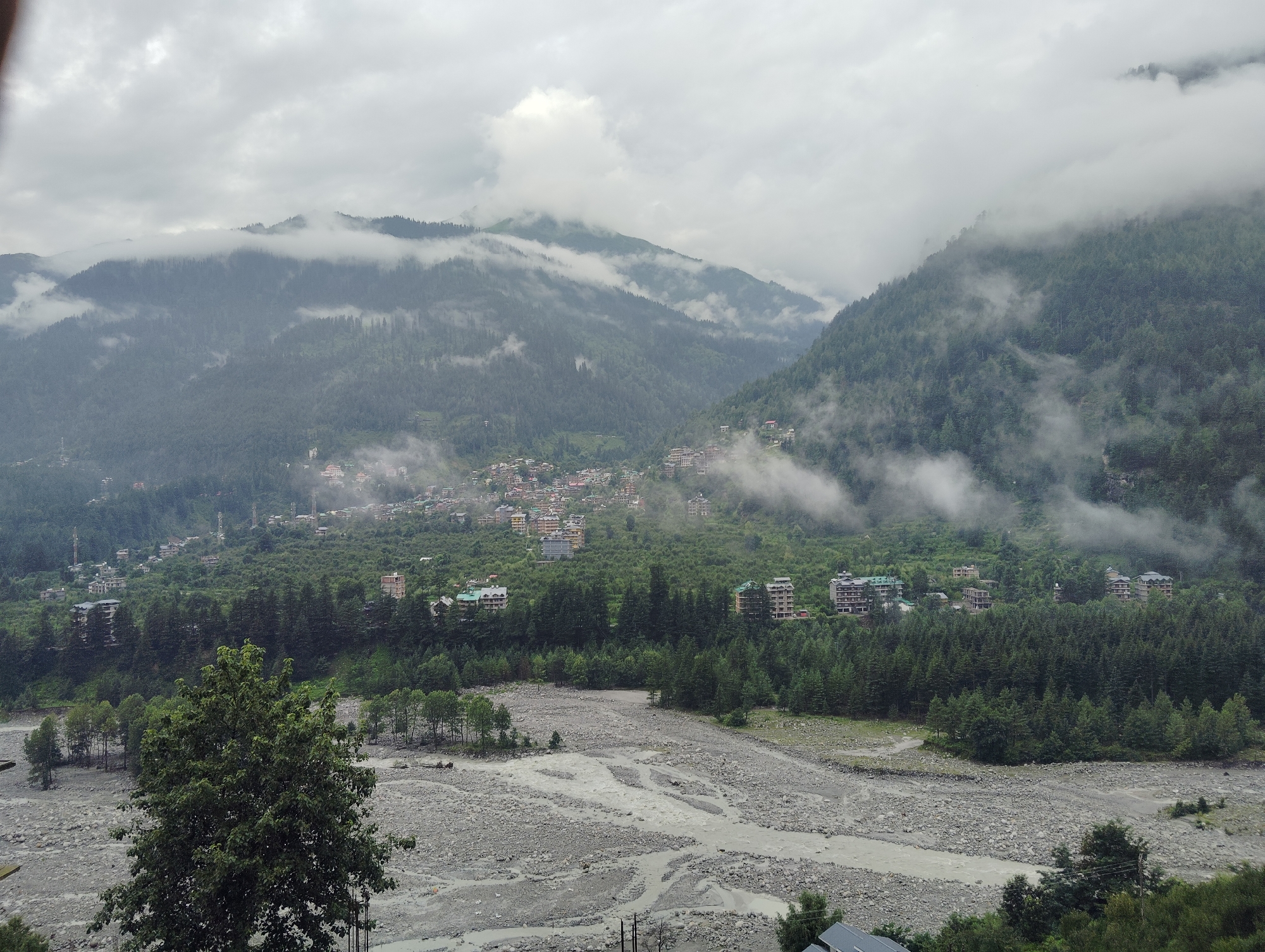 The image captures a mountainous landscape with a village nestled in the valley. The sky is overcast with thick clouds, creating a moody atmosphere. A riverbed filled with grey stones and a small stream flows in the foreground. The middle ground features a dense forest of pine trees, with buildings of the town of Vashisht, India, scattered across the lower slopes of the mountains. The town is characterized by a mix of colorful buildings and modern structures, some with multi-story architecture. The upper slopes of the mountains are covered in lush green vegetation and further obscured by mist and clouds. The overall impression is one of a serene and remote mountainous region experiencing cloudy weather.