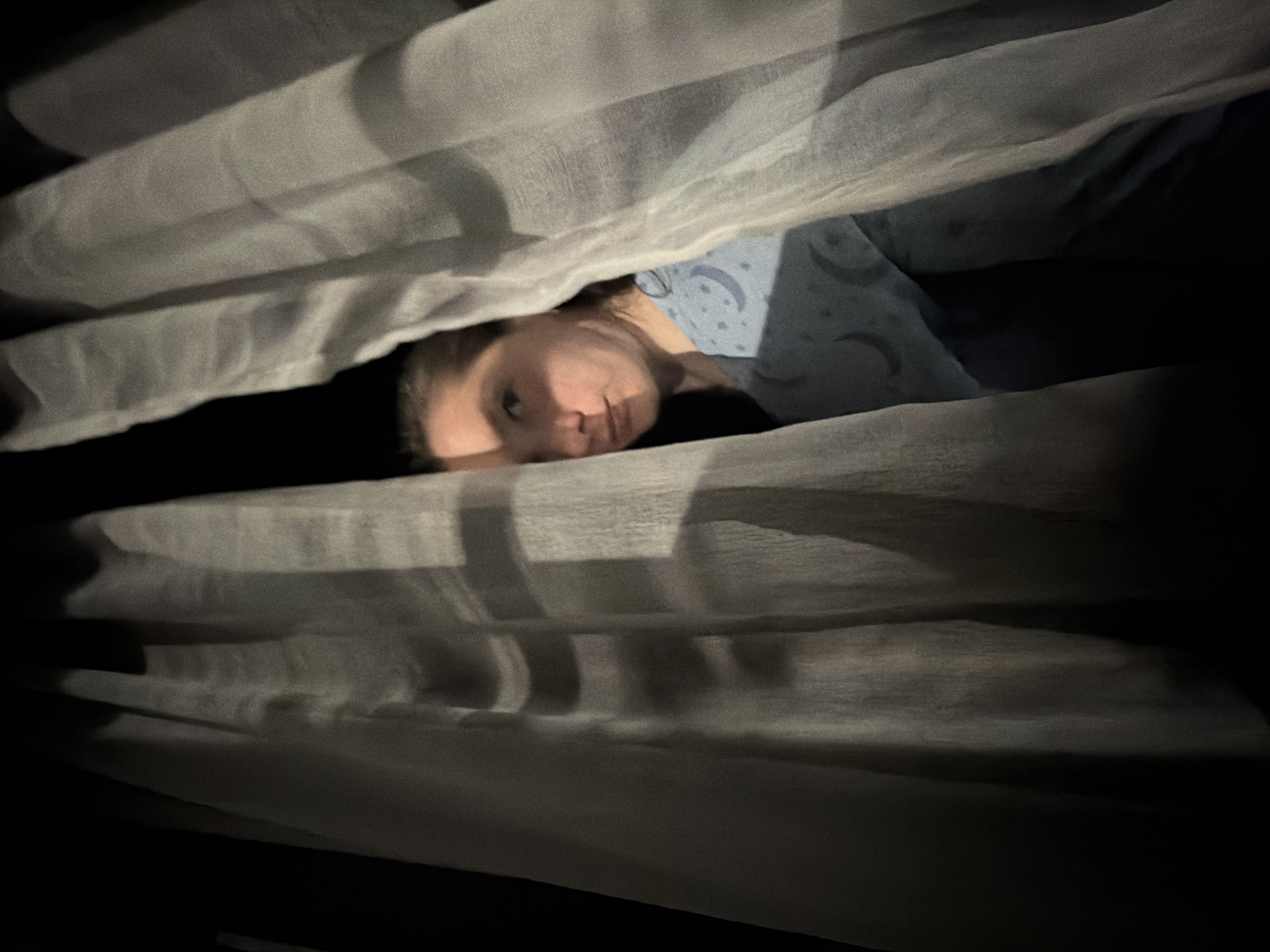A person is lying in bed under white sheets. The person appears to be female with fair skin, and is wearing a blue top with a moon and star pattern. The environment is dimly lit, with a dark background. The image's location is not discernible.
