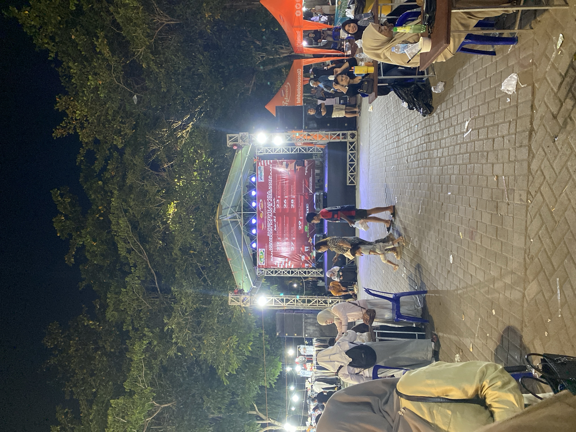 The image depicts an outdoor event at night, likely a fair or festival, set in the city of Banjarmasin, Indonesia. In the background, a large stage is illuminated, featuring a red banner with text that appears to be promotional or informational, possibly related to an event or advertisement. Several individuals are present, some are seated at tables, and others are standing and walking around. The ground is paved with interlocking bricks, and there are trees with dense foliage overhead, suggesting a park or open-air venue. Scattered litter is visible on the ground, indicating a public gathering. In the foreground, several people are seen from the back, some wearing hijabs, interacting with each other or moving through the space. The overall atmosphere suggests a lively evening event.