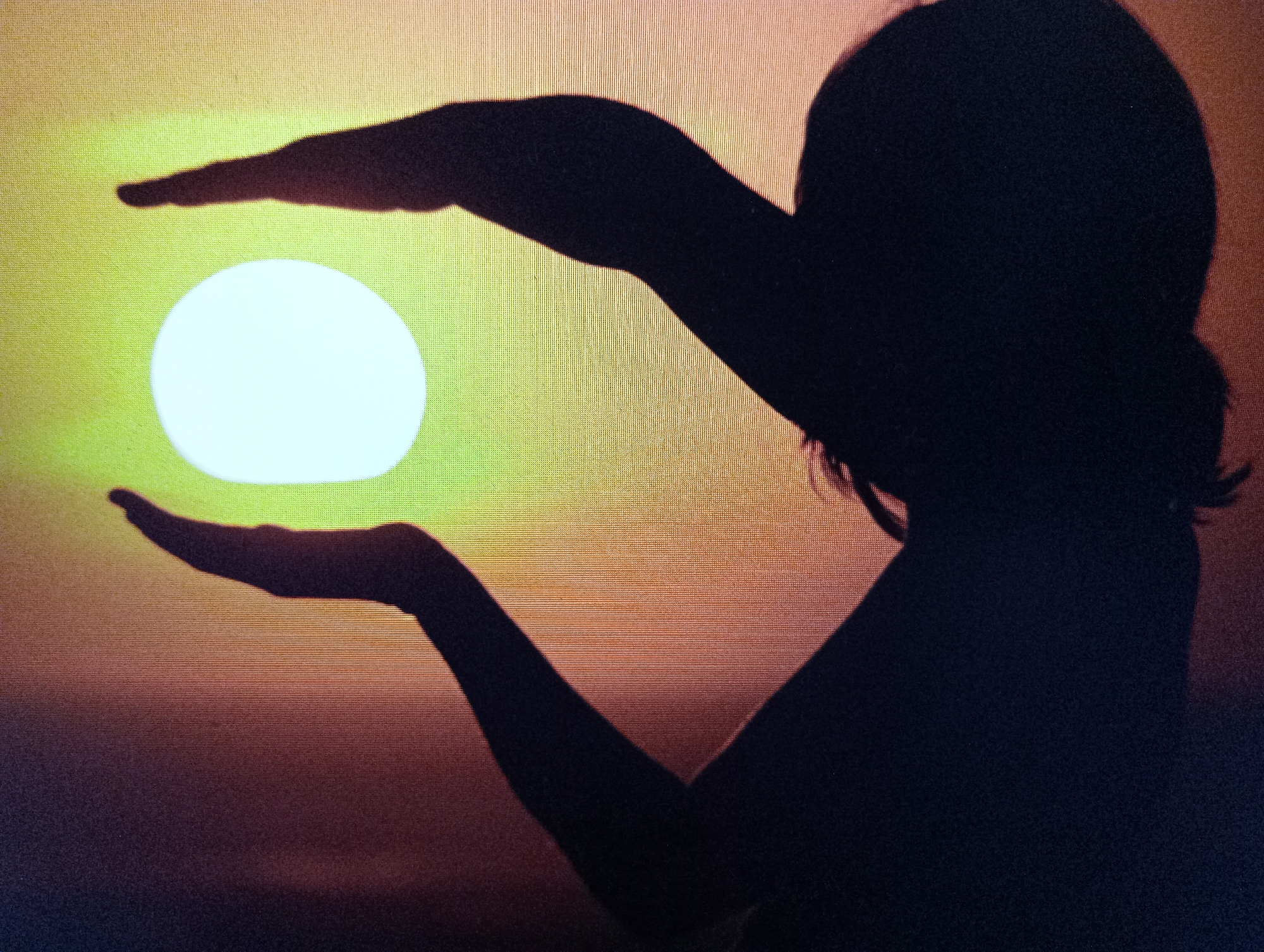 A silhouette of a person's head and arms is framed against a bright, yellowish-green sun, which appears to be setting. The sun is a circular shape, held between the person's cupped hands. The person's body is also in silhouette, appearing to be facing the right side of the frame. The background is a gradient of colors, transitioning from yellow-green at the top to darker hues of orange and purple towards the bottom. This image was taken at the Harmanli, Bulgaria location.