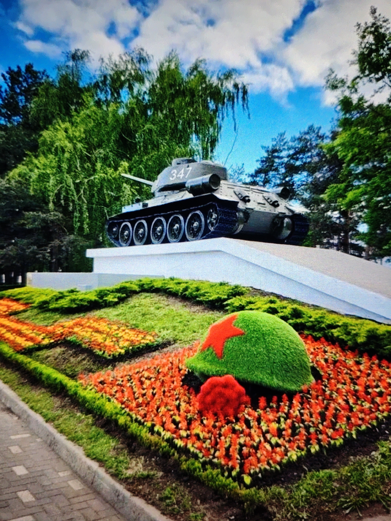 The image depicts a monument featuring a Soviet-era tank, identified by the number "347" on its turret, positioned on a white pedestal. The tank is a T-34 model. In the foreground, there is a meticulously arranged flower bed with vibrant orange and yellow flowers, bordered by green grass. A spherical topiary adorned with a red star and a cluster of red flowers sits within the flower bed. The background consists of lush green trees and a bright blue sky with scattered white clouds. This monument is located at the Memorial Complex "Eternal Glory" (Pamyatnik "Vechnaya Slava") in Stavropol, Russia, dedicated to fallen soldiers.