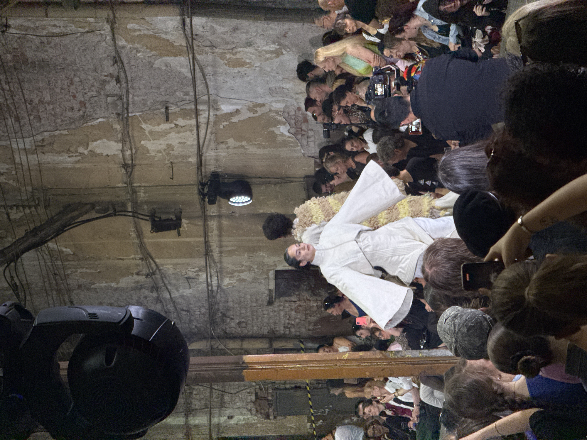 This image captures a fashion show taking place in an industrial-style venue in Sofia, Bulgaria. The audience, composed of a diverse group of people, is seated on the left and right sides of a runway. The runway itself is bare concrete. On the runway, a model in an avant-garde white outfit with voluminous sleeves is walking towards the viewer. Another model, partially visible behind the first, is wearing a textured, pale yellow and white garment. The background of the venue features distressed, textured walls with visible brickwork, and industrial lighting fixtures, including a large, black spotlight hanging from the ceiling. Various cameras and recording devices are being used by attendees to document the event. The overall atmosphere suggests a contemporary and edgy fashion presentation.