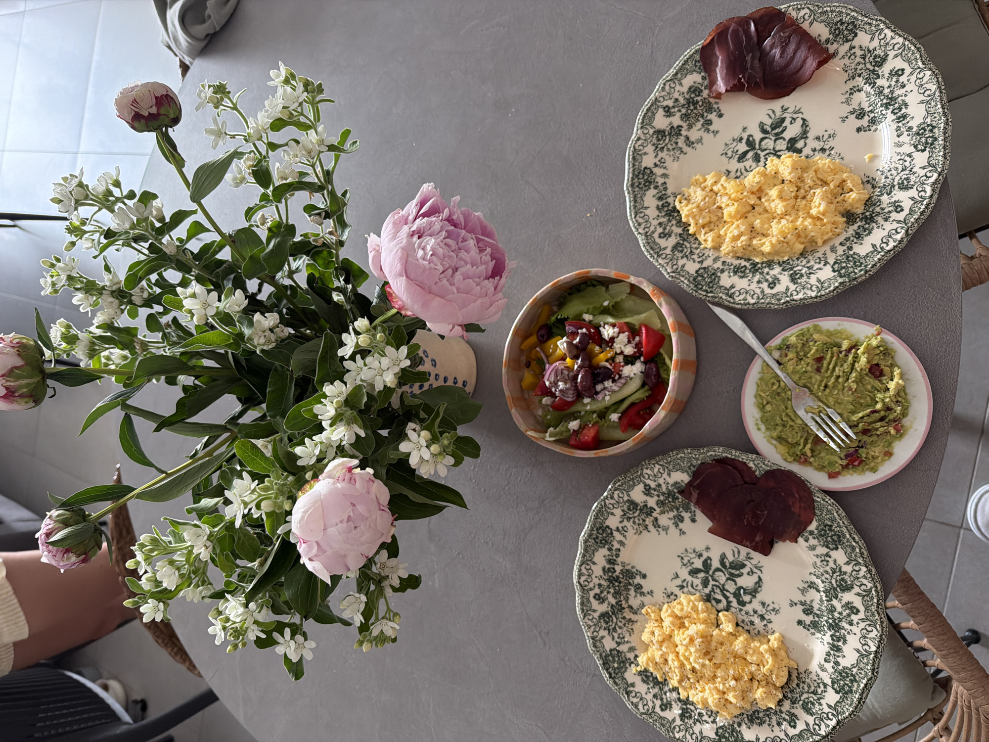 The image depicts a grey round table set with a vase of flowers and various plates of food. The floral arrangement includes pink peonies and small white flowers. Plates of food include scrambled eggs with what appears to be bresaola, a salad in a striped bowl, and a small plate with guacamole and a fork. The setting gives the impression of a meal being prepared or eaten at home.