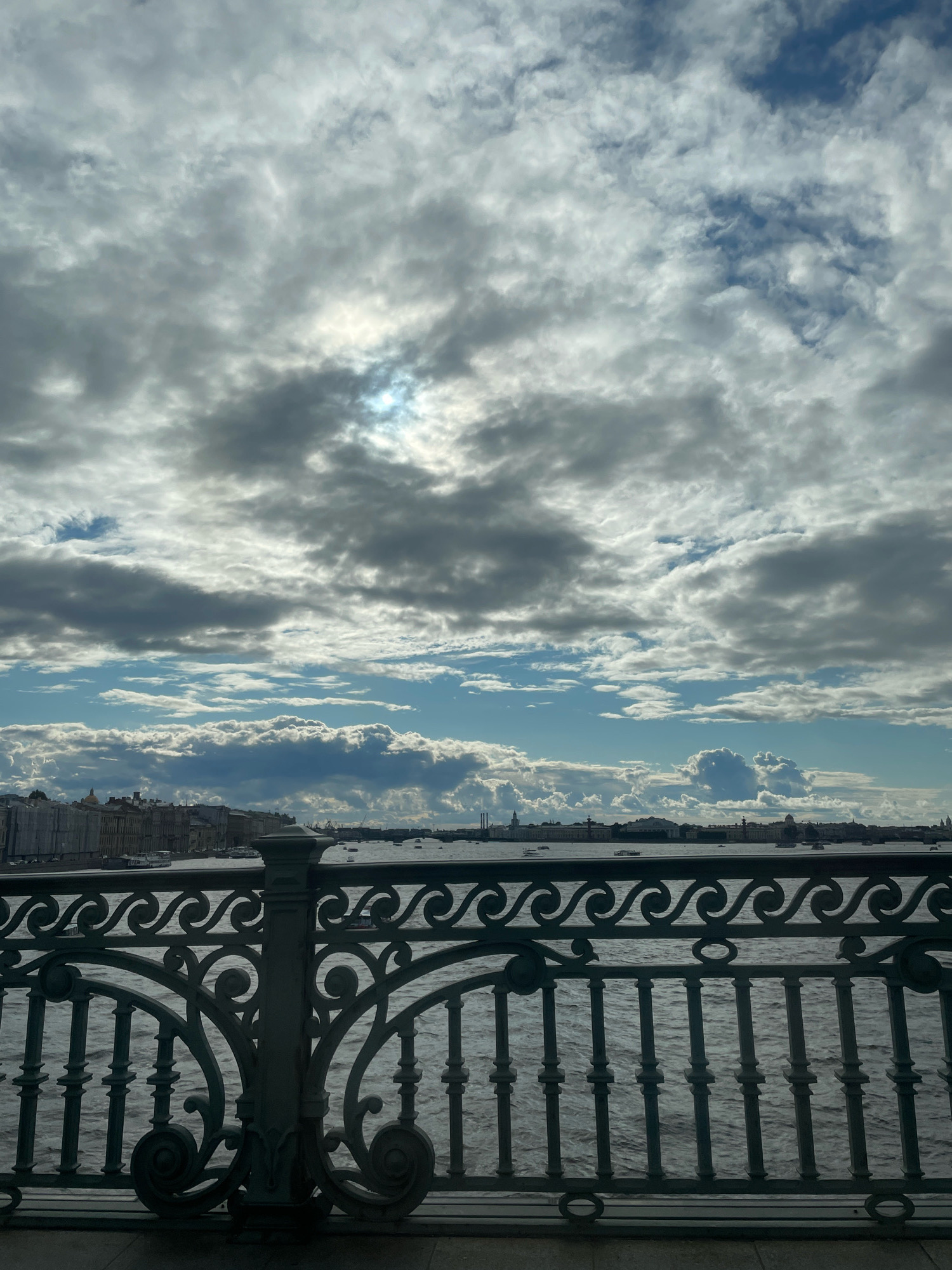 This is a photograph taken from a bridge over a body of water, likely a river, in Saint Petersburg, Russia. In the foreground, a decorative metal railing with intricate scrollwork and vertical bars separates the viewer from the water. The water itself appears choppy with a moderate ripple. Beyond the water, a cityscape is visible, featuring a line of buildings with various architectural styles, some appearing to be under construction or renovation. Several boats are present on the water. The sky above is dramatic, filled with a mixture of dark, heavy clouds and brighter, scattered clouds, with glimpses of blue sky and the sun peeking through in the upper center of the frame. The overall lighting suggests it is daytime, but overcast with breaks of sunshine.
