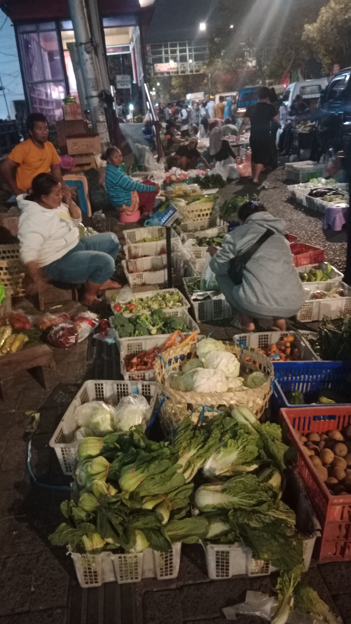 The image depicts an outdoor market at night in Semarang, Indonesia. A group of people, likely vendors and customers, are gathered around stalls displaying a variety of fresh produce. Numerous white plastic crates and baskets are filled with vegetables such as cabbage, broccoli, carrots, and leafy greens. Some individuals are seated, appearing to be vendors attending to their goods, while others are standing and browsing. In the background, several vehicles, including cars and a van, are visible, suggesting the market is located near a road. The scene is illuminated by artificial lighting, creating a dynamic atmosphere typical of a bustling night market.
