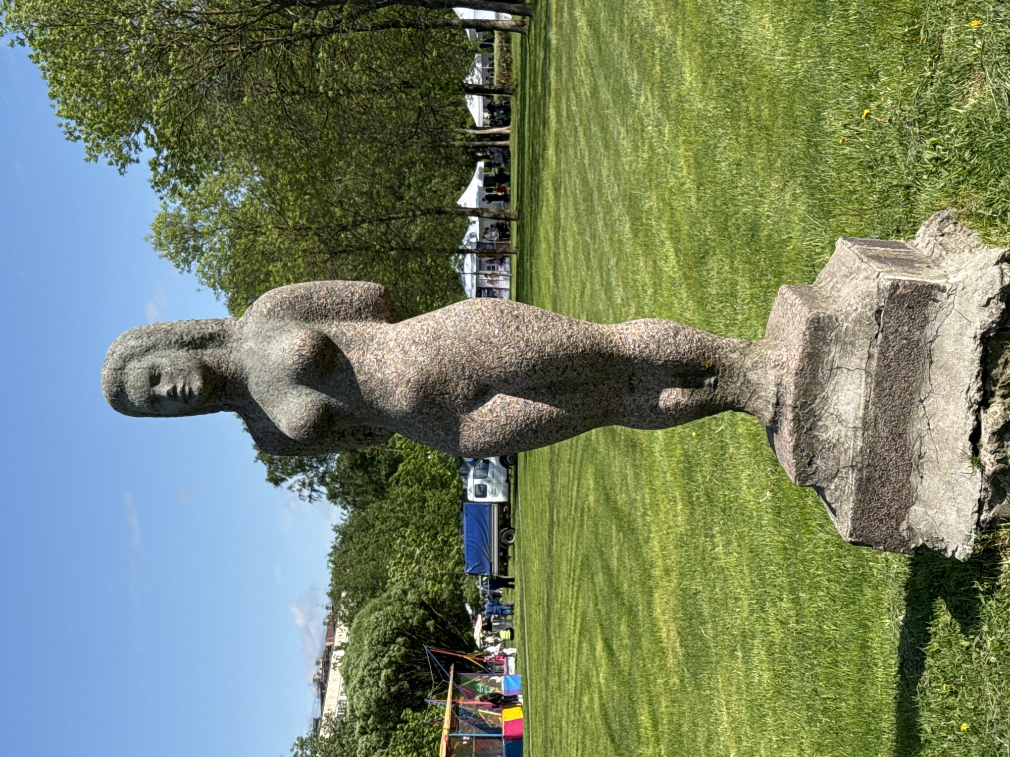 Here is a description of the image:
A full-length statue of a nude female figure stands in a grassy park on a sunny day. The statue is made of textured stone and is mounted on a stone pedestal. In the background, there is a grassy field, a line of trees, and a blue sky. Further back, there are tents, a truck, and a colorful play structure. The image was likely taken in Saint Petersburg, Russia, based on the style of the buildings and the presence of the statue in a public park setting.
