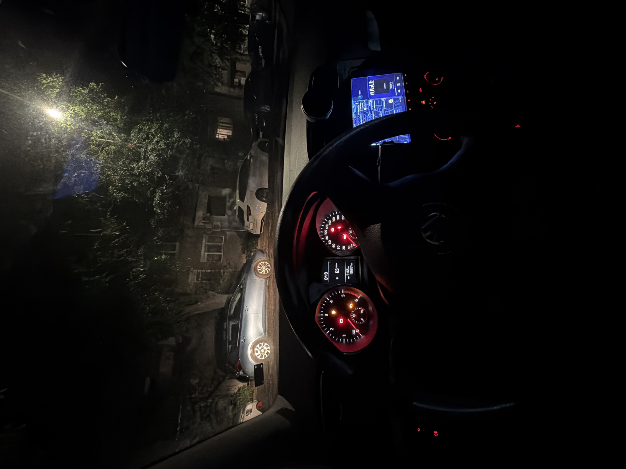 The image depicts a view from inside a parked car at night. The car's dashboard is visible, with illuminated gauges and a navigation screen displaying a map. Through the windshield, a street scene is visible, showing parked cars along the curb and a building illuminated by a streetlight. The buildings suggest an urban environment. While there is no confirmation that it is in the city of Burgas, Bulgaria. There are no people visible in the image.