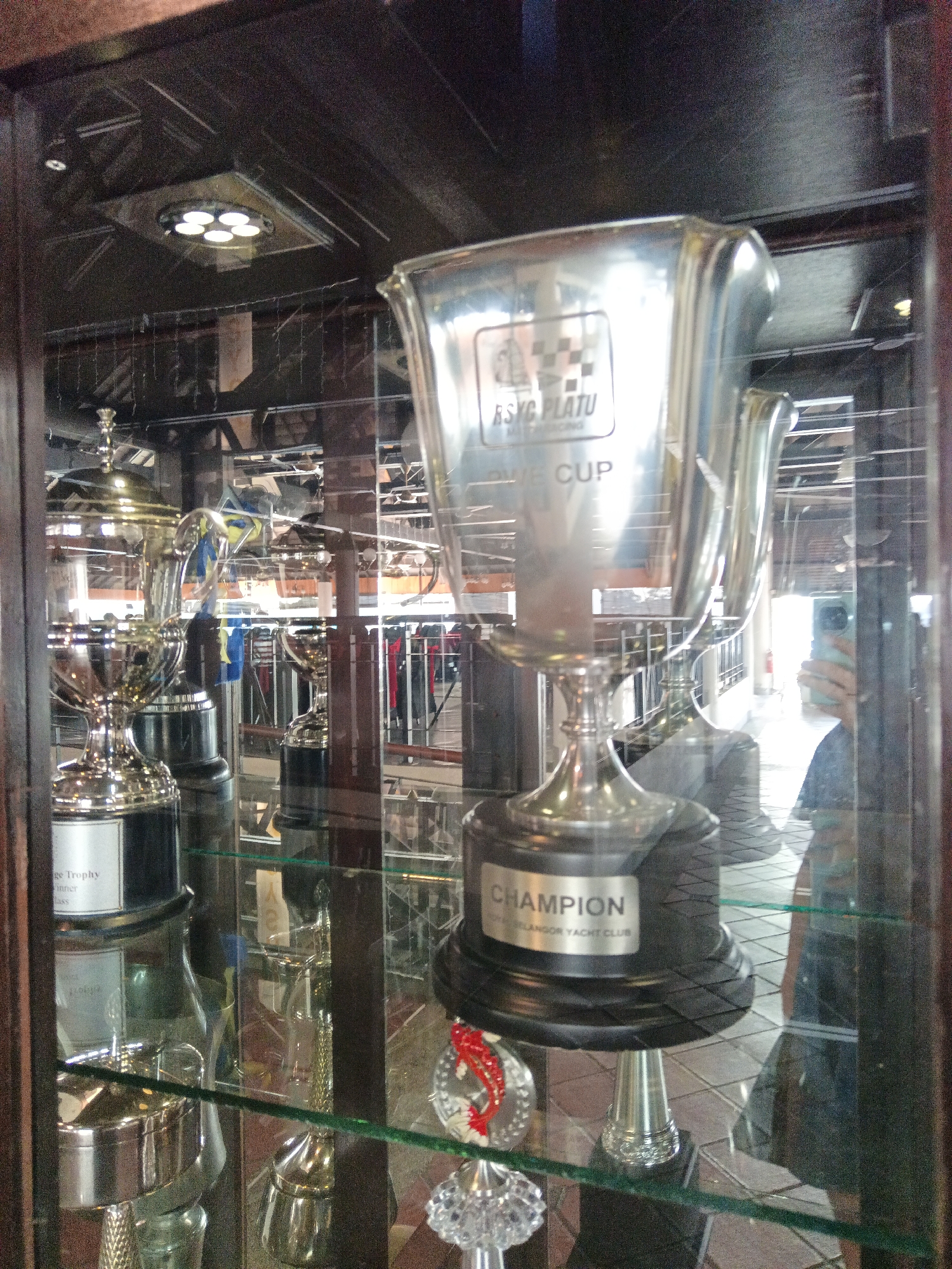 The image shows a display case containing several trophies, the most prominent of which is a large silver cup with "CHAMPION" and "SELANGOR YACHT CLUB" inscribed on its base. The trophy is positioned in the center of the frame, reflecting the surrounding environment. Other trophies of varying sizes and designs are visible to the left and right. The display case appears to be made of glass and wood, and is illuminated by overhead lights, one of which is visible at the top of the image. The background shows an indoor setting with what appears to be a public area, possibly a club or building lobby, with a person's reflection visible in the glass of the display case on the right side. The overall setting suggests a location where sporting achievements are celebrated. This location is identified as the Selangor Yacht Club in Klang City, Malaysia.