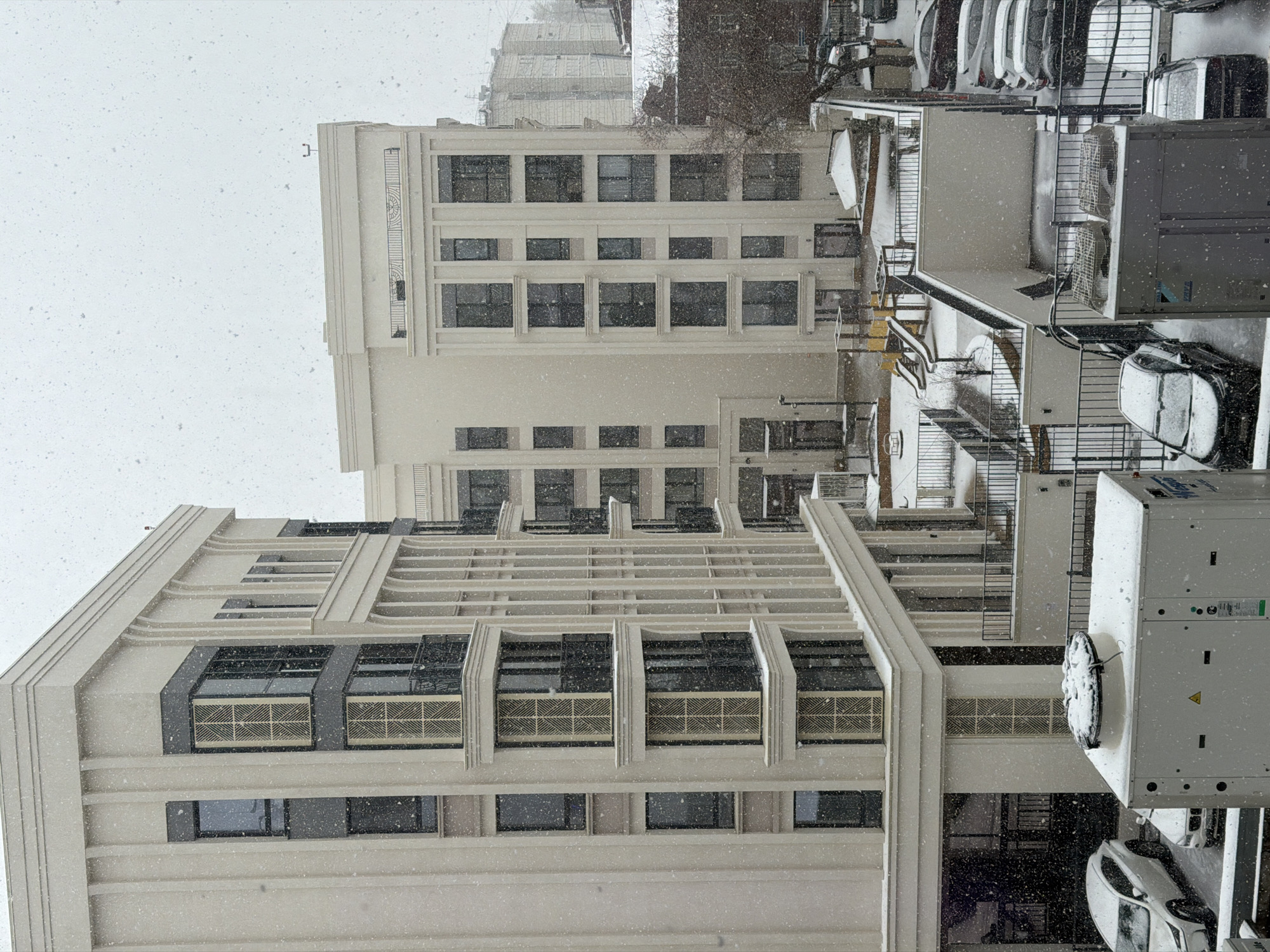 This image presents an urban landscape in Perm, Russia, engulfed in a heavy snowfall. Two prominent multi-story buildings with light-colored, possibly beige or off-white, facades dominate the scene. Their architecture features strong vertical and horizontal lines, with numerous rectangular windows, some showcasing decorative grilles or vents with intricate geometric patterns below them.
The ground level and surrounding areas reveal a parking lot or street with numerous cars, many visibly covered in a fresh layer of snow, attesting to the ongoing heavy precipitation. Several large, box-like outdoor units, likely HVAC equipment, are situated on rooftops or platforms, also accumulating snow. Metal railings line balconies, stairways, and other elevated structures.
The sky is a uniformly overcast white, filled with countless snowflakes sharply visible in the air, creating a dense, wintry atmosphere. The photograph is taken from an elevated perspective, looking down into a courtyard or street. No people are visible, suggesting the intense cold and snow have kept activities indoors. A small, C-shaped fixture is noticeable on the top edge of the closer building. On one large white utility unit, a yellow triangular warning symbol is present, but no other text is clearly legible. The overall impression is one of serene, cold tranquility, with the fresh snow transforming the urban environment into a monochromatic winterscape.