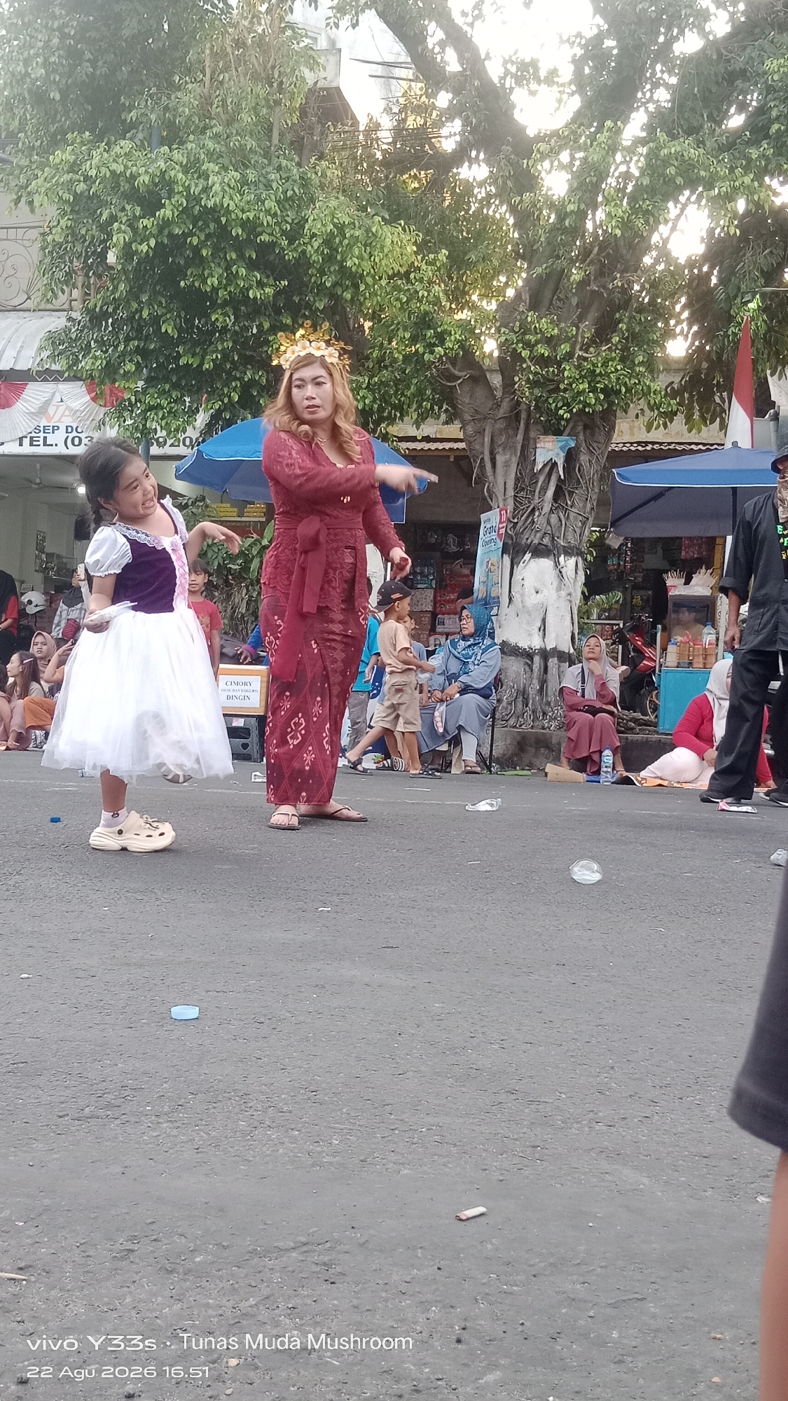 In Genengan, Indonesia, a street performance is underway featuring a young girl in a white dress and a woman in a traditional red outfit with a gold headpiece. The girl stands to the left, facing the viewer with her mouth open and her right arm extended as if gesturing or singing. She wears a white dress with purple accents and tan slip-on shoes. To her right, the woman is in profile, pointing her right index finger forward. She wears a long, red patterned garment tied with a sash. Behind them, spectators are seated and standing. To the left, a blue umbrella is visible. The ground is asphalt, with some scattered debris including a clear plastic bottle on the left and a white object on the right. A large tree with dense green foliage dominates the background, with a hint of buildings and market stalls behind it. The lighting suggests it is daytime, possibly late afternoon.